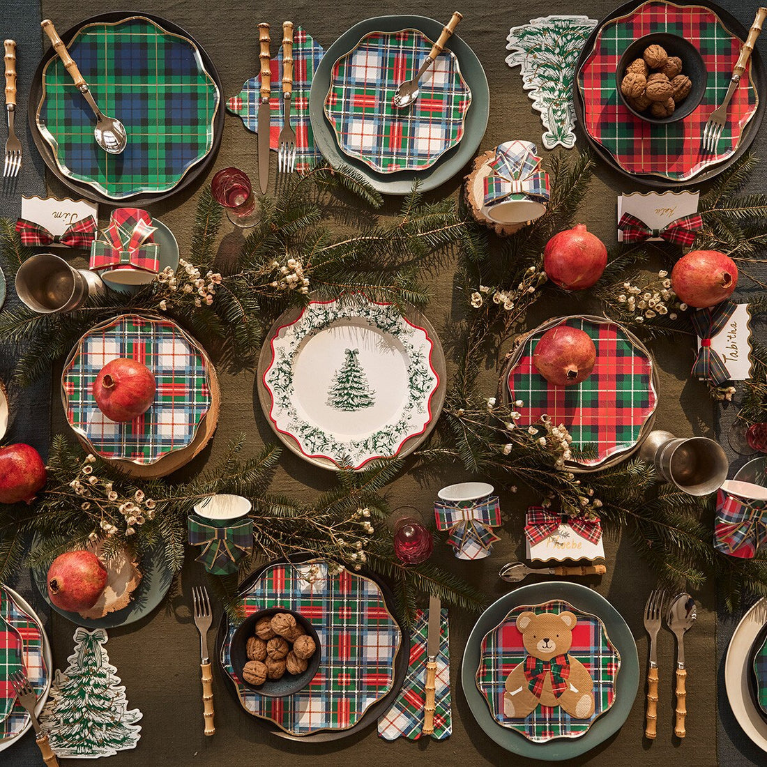 A festive holiday table setting featuring multiple green, red, and white plaid placemats, matching napkins, green ceramic plates, wooden cutlery, pine garlands, pomegranates, and small decorative Christmas tree cutouts on a dark wooden table.
