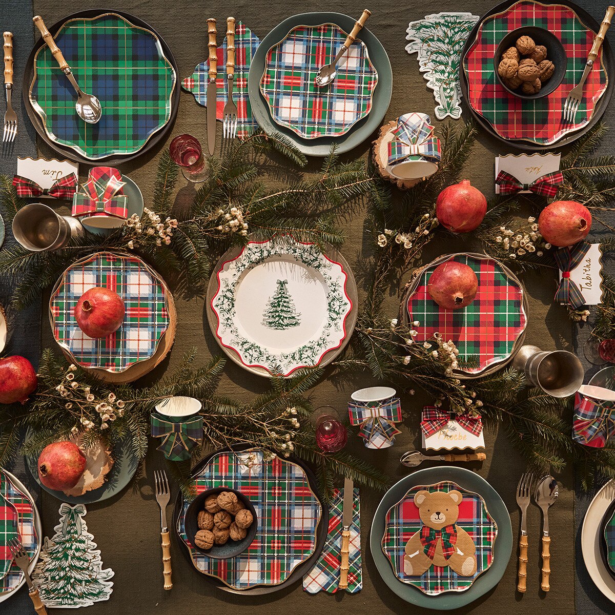 A festive holiday table setting featuring multiple green, red, and white plaid placemats, matching napkins, green ceramic plates, wooden cutlery, pine garlands, pomegranates, and small decorative Christmas tree cutouts on a dark wooden table.