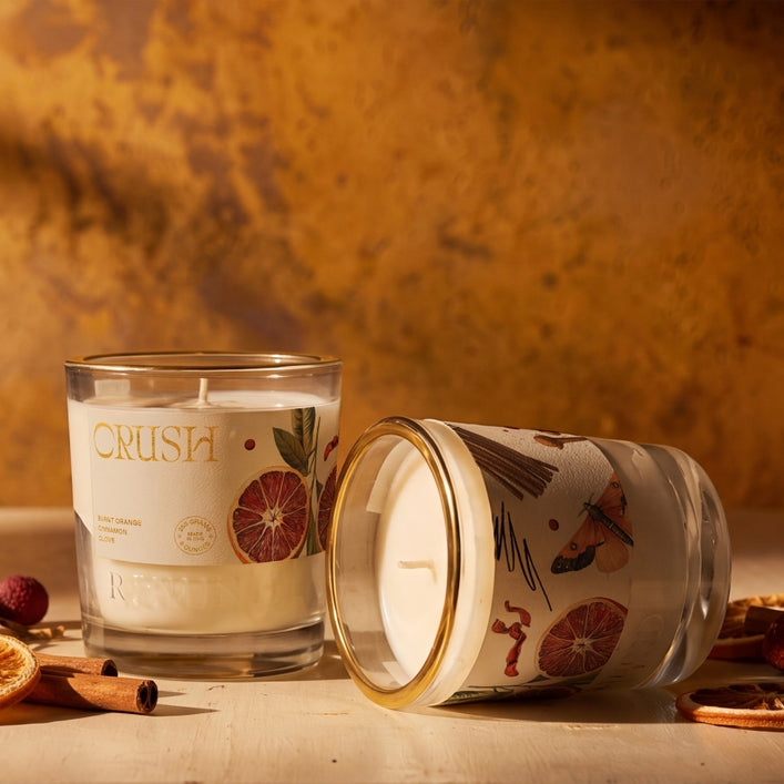 Two scented candles in clear glass jars with gold rims, one standing upright and one lying on its side, set on a tabletop with dried citrus slices, cinnamon sticks, and a warm textured background. The upright jar shows the brand name CRUSH and a citrus slice illustration on the label.