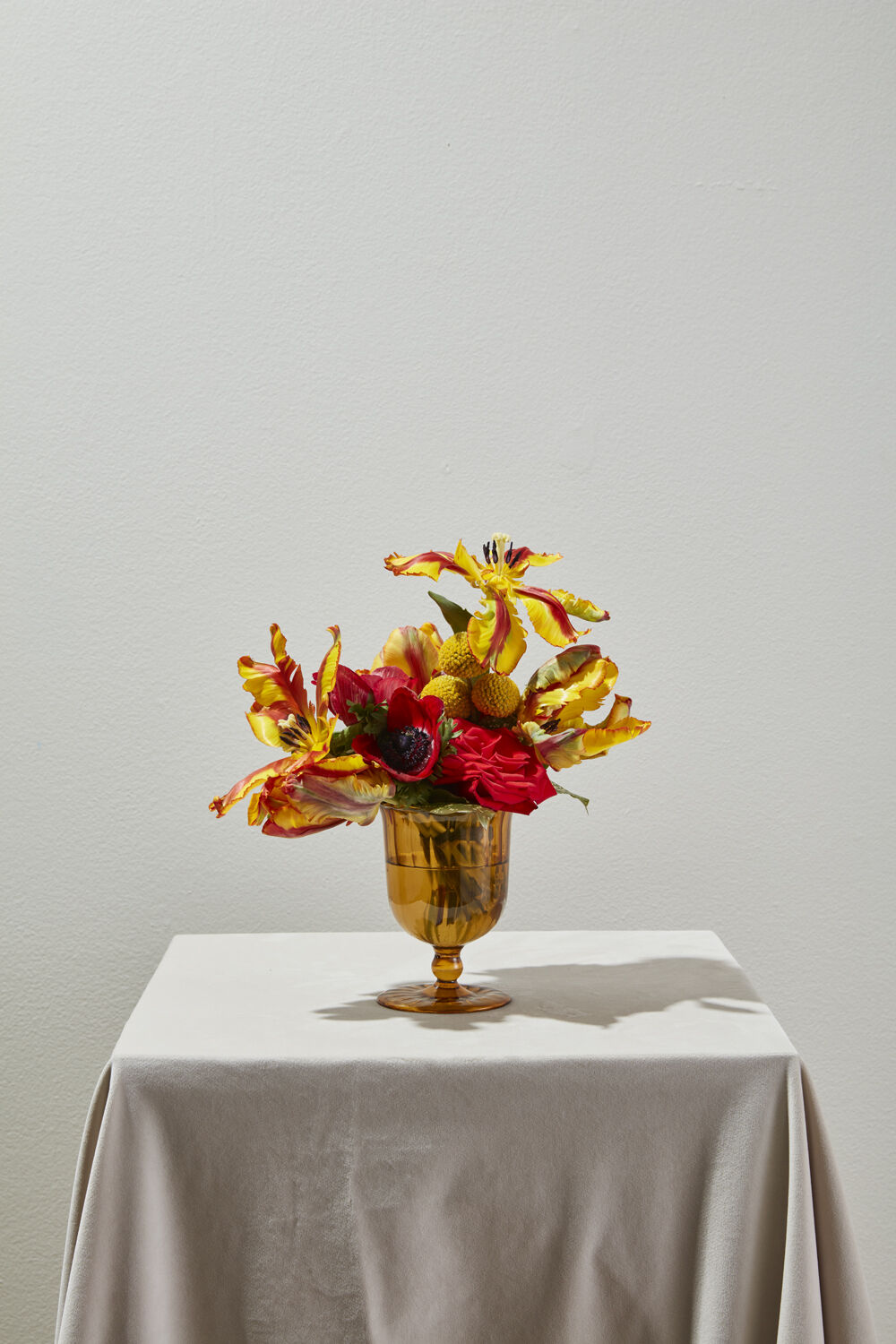 A decorative floral arrangement with yellow, red, and orange flowers in a gold-colored glass vase, displayed on a draped white table against a plain light gray wall.