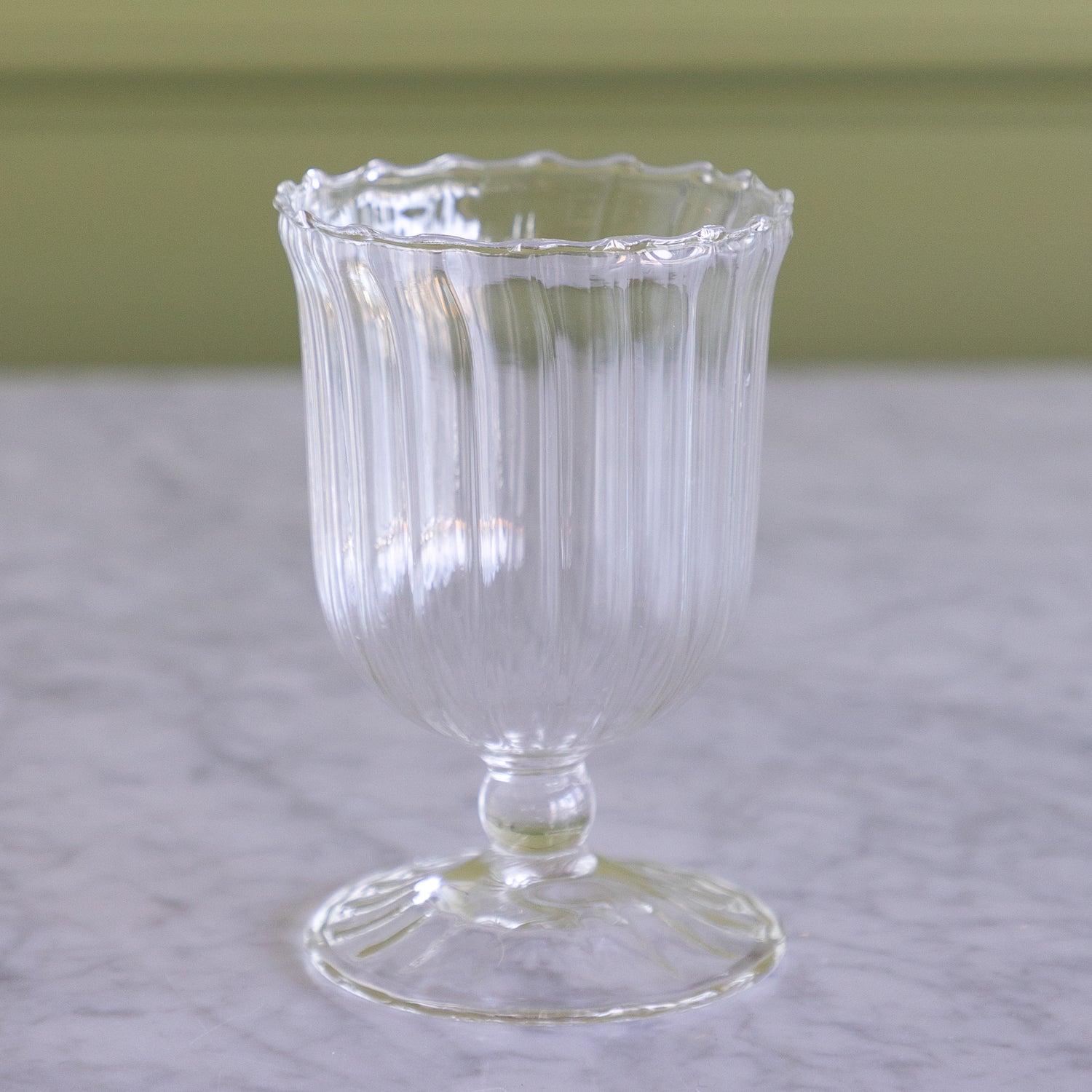 A clear ribbed glass cup with a scalloped rim, stem, and pedestal base, shown on a light marble surface with a soft olive-green background.