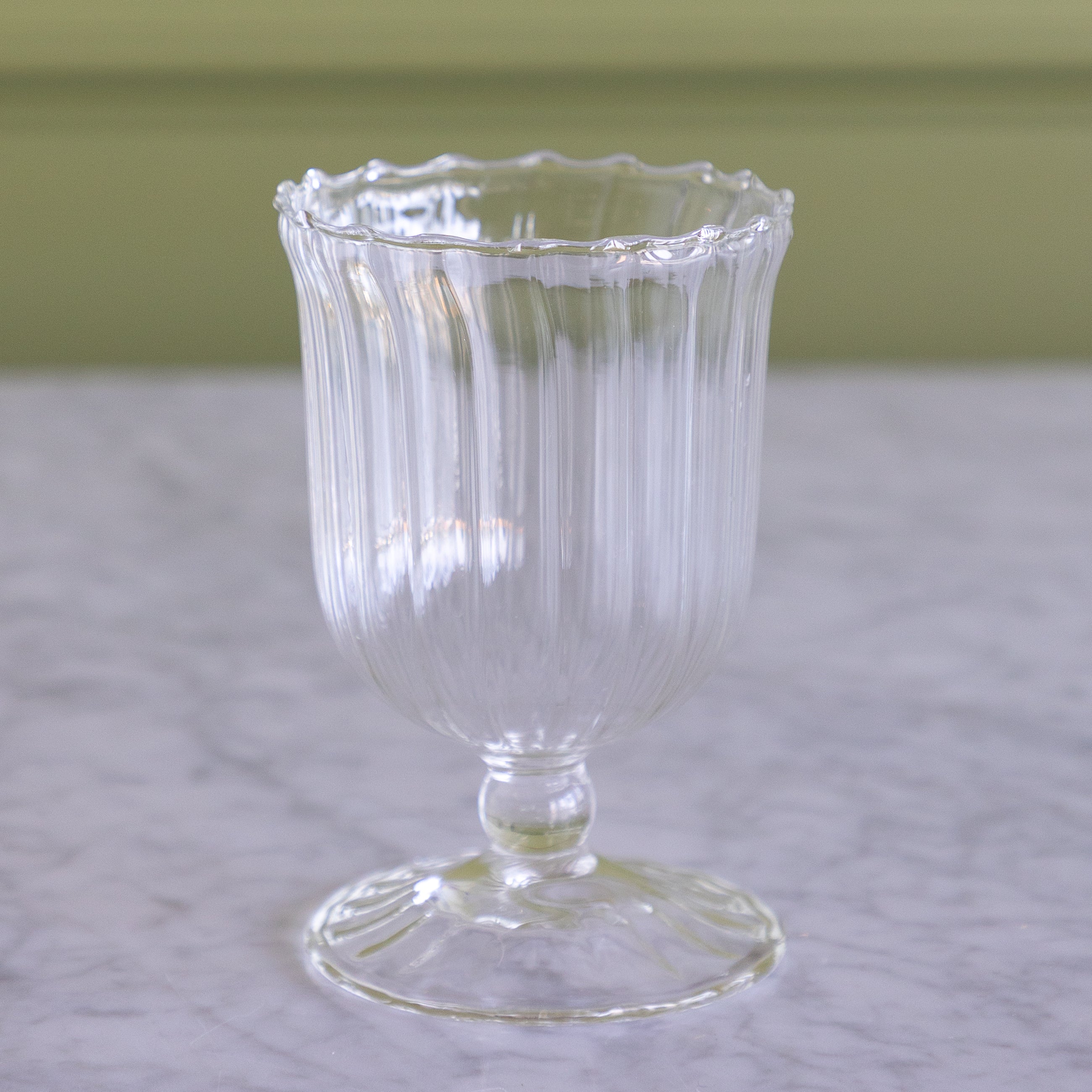 A clear ribbed glass cup with a scalloped rim, stem, and pedestal base, shown on a light marble surface with a soft olive-green background.