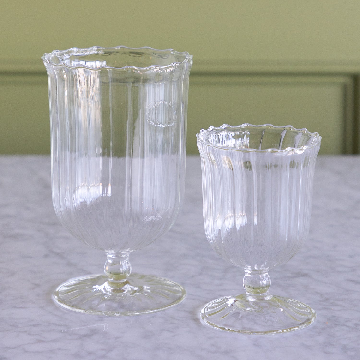 Two clear ribbed glass goblets, one tall and one short, displayed on a light marble surface against a soft olive-green paneled background.