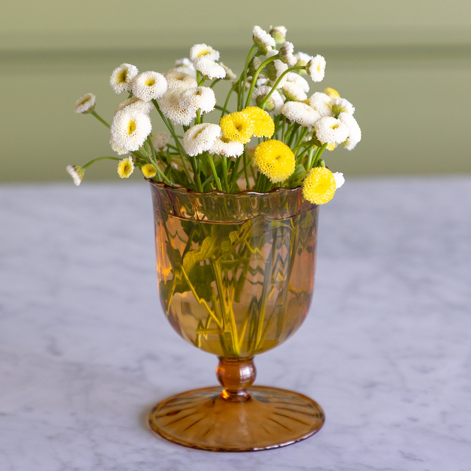 An amber glass goblet vase filled with white and yellow daisy-like flowers, set on a light marble surface against a soft olive-green background.
