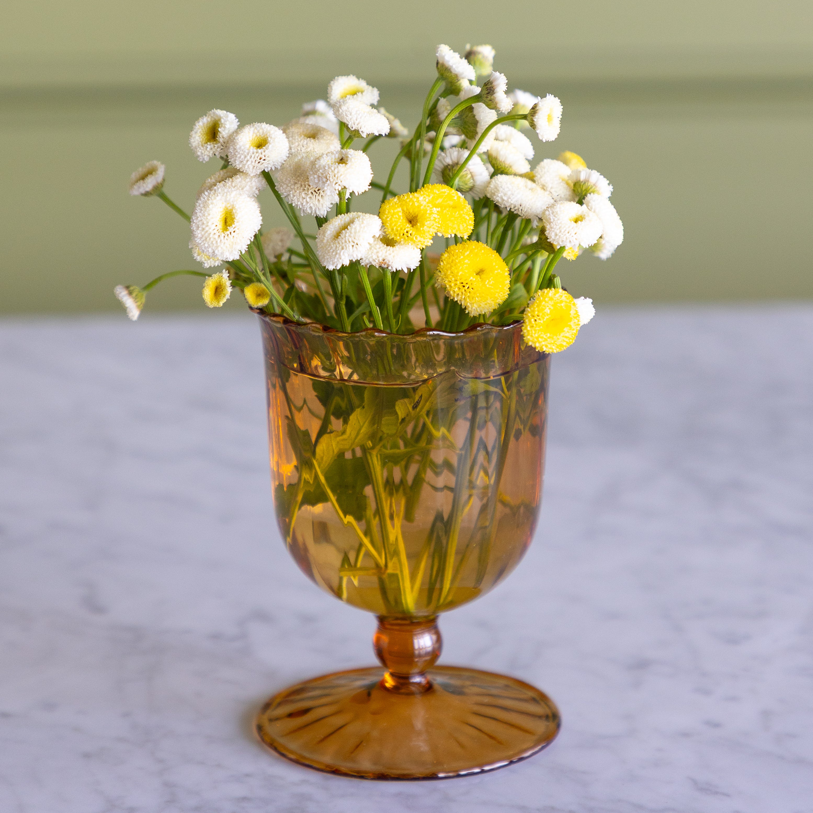 An amber glass goblet vase filled with white and yellow daisy-like flowers, set on a light marble surface against a soft olive-green background.