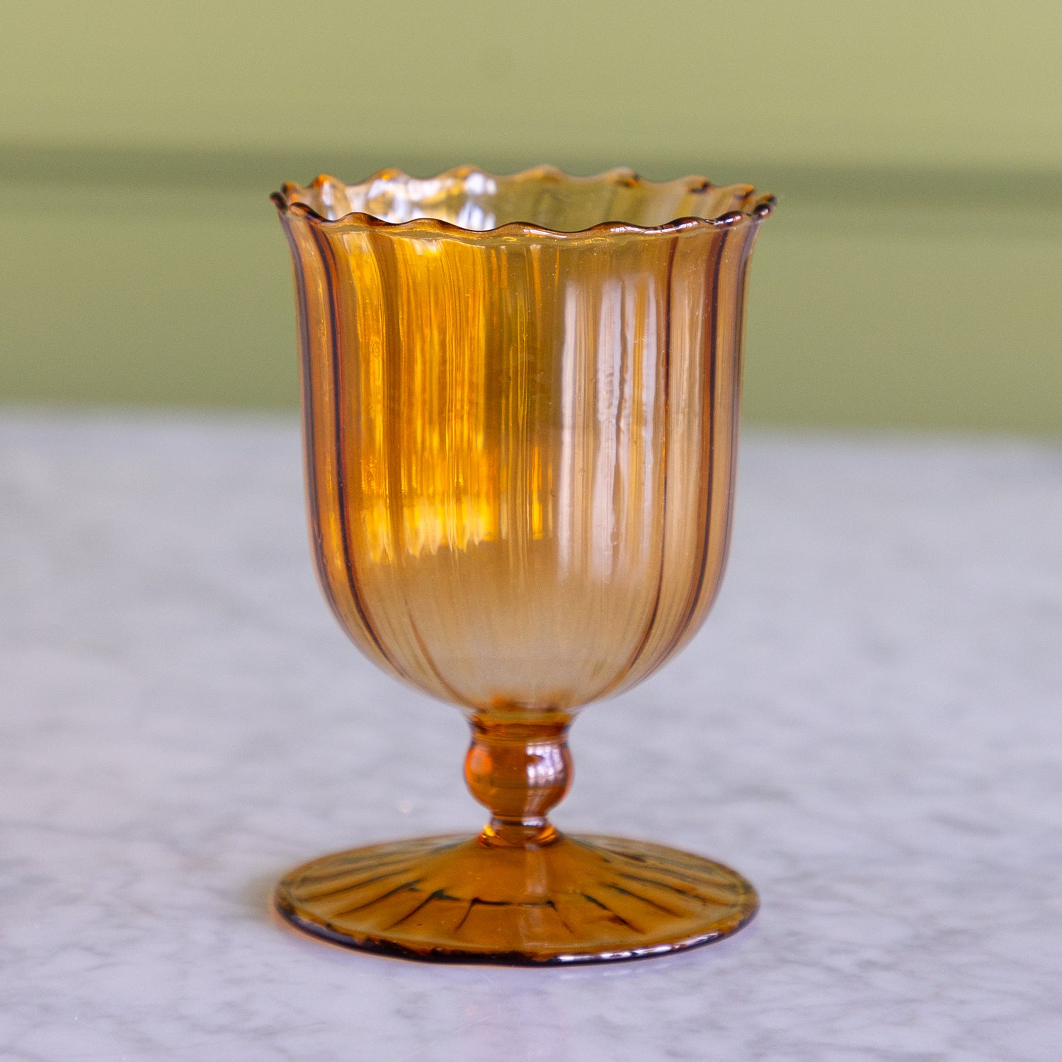 An amber-colored ribbed glass goblet with a scalloped rim and pedestal base, standing on a light marble surface against a soft olive green background.