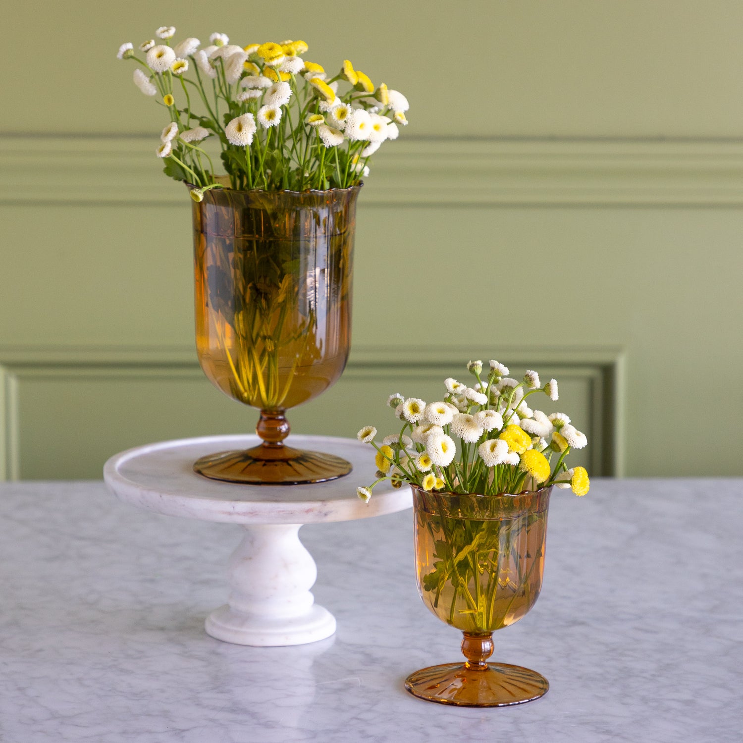 Two amber glass goblet vases filled with white and yellow daisies, displayed on a white marble pedestal and tabletop against a sage green paneled wall.