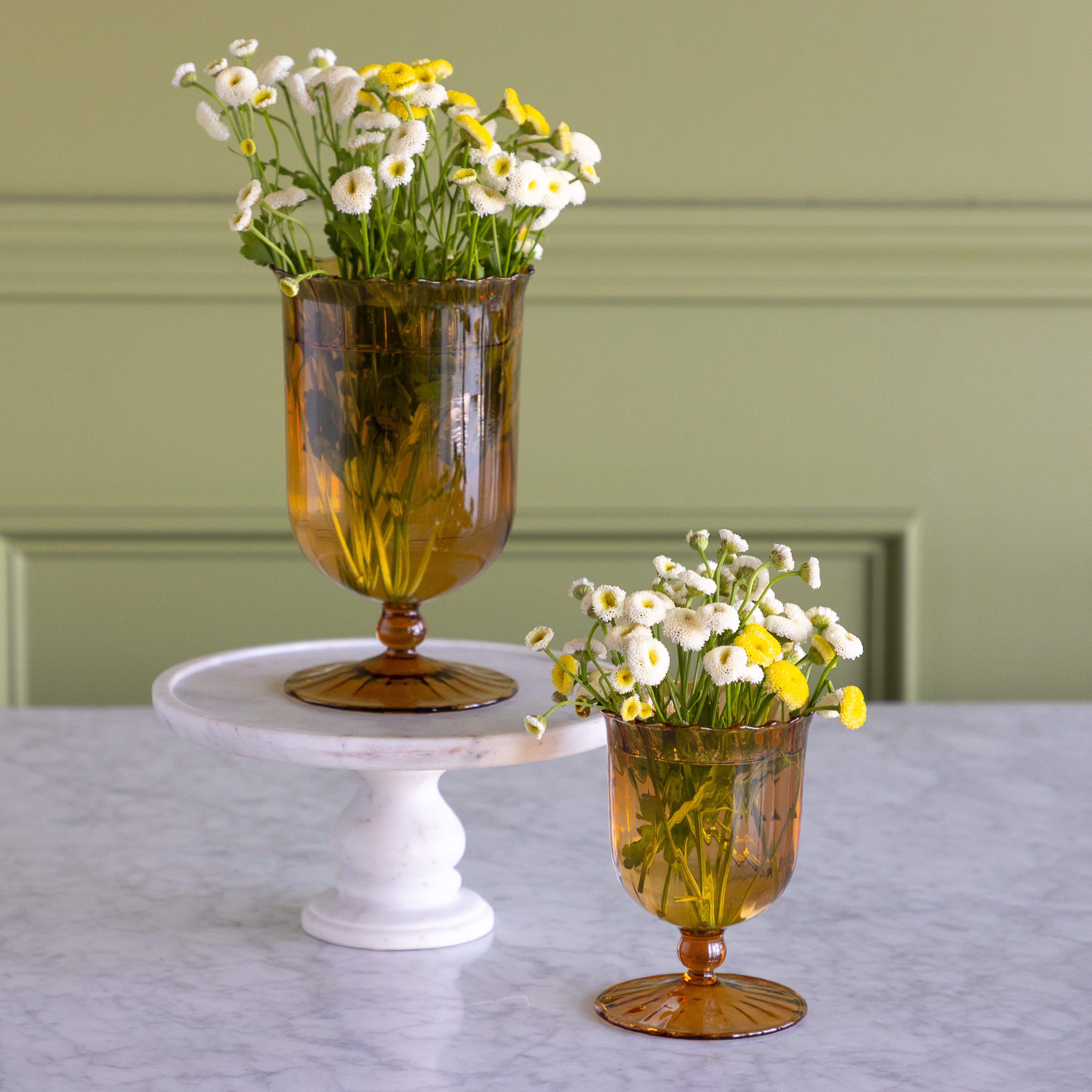 Two amber glass goblet vases filled with white and yellow daisies, displayed on a white marble pedestal and tabletop against a sage green paneled wall.