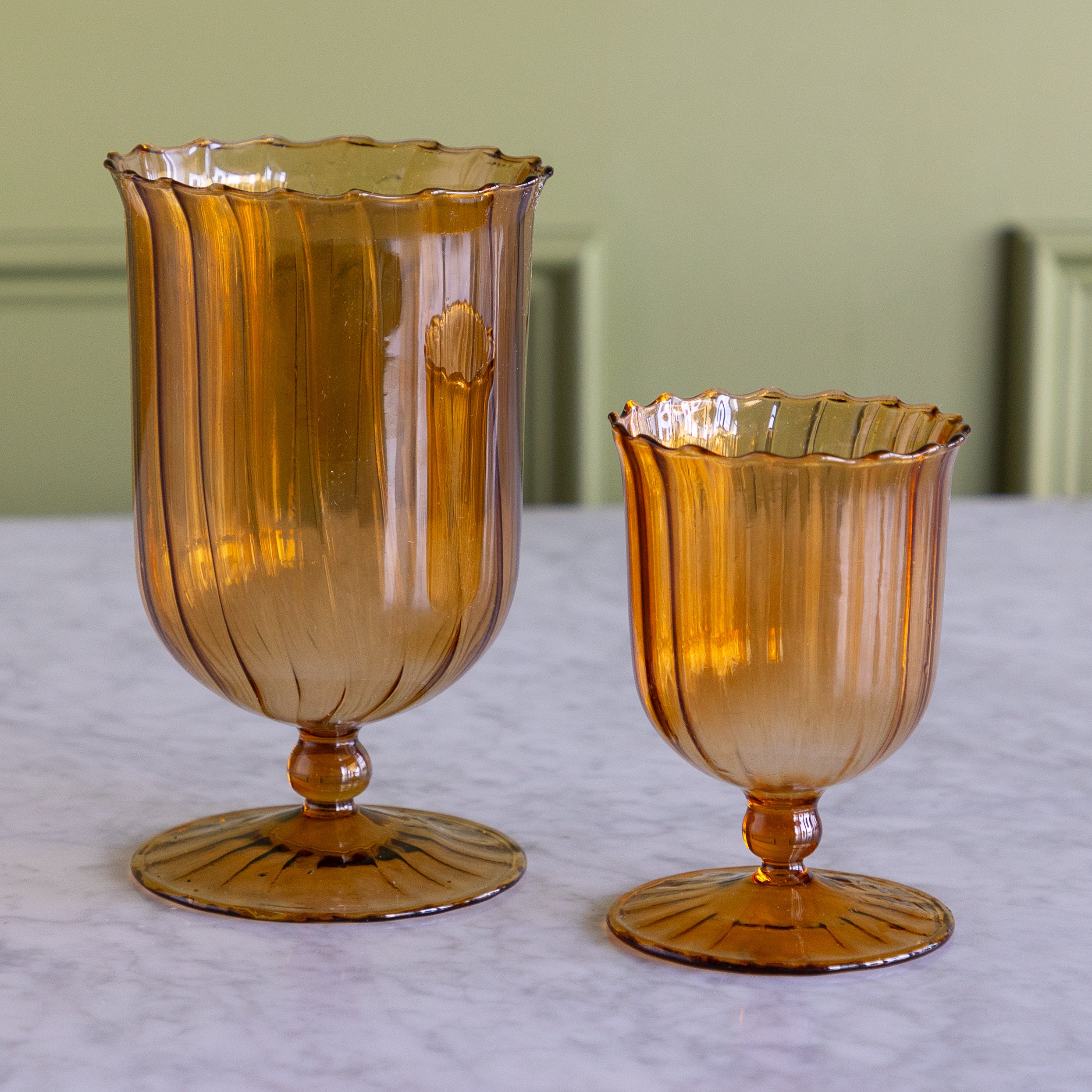 Two amber glass pedestal vases of different sizes on a light marble surface with a soft-focus green wall background.