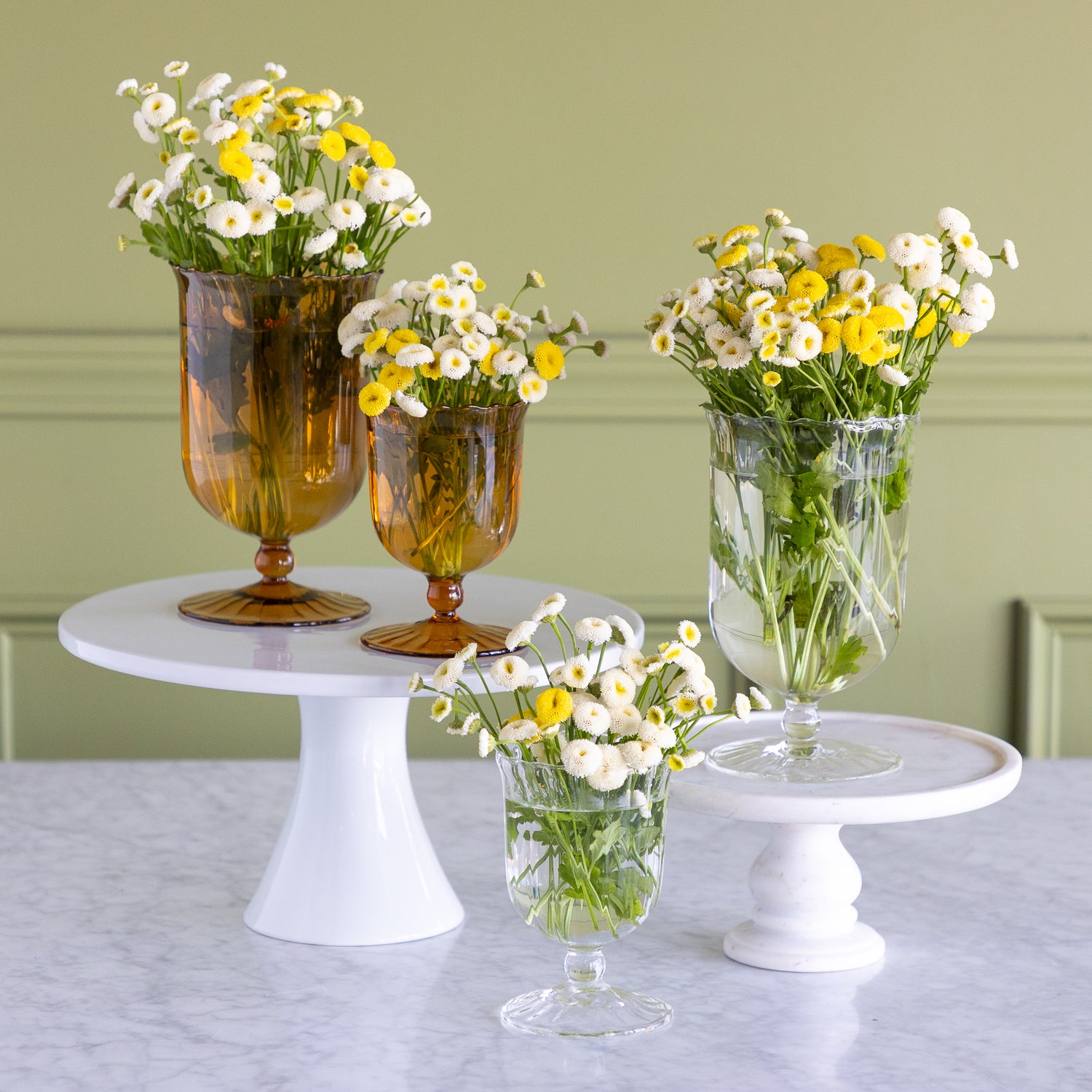 Two amber glass vase holders and two clear glass vase holders arranged on white pedestal stands with white and yellow daisy flower arrangements, set against a sage green paneled wall and light marble tabletop.