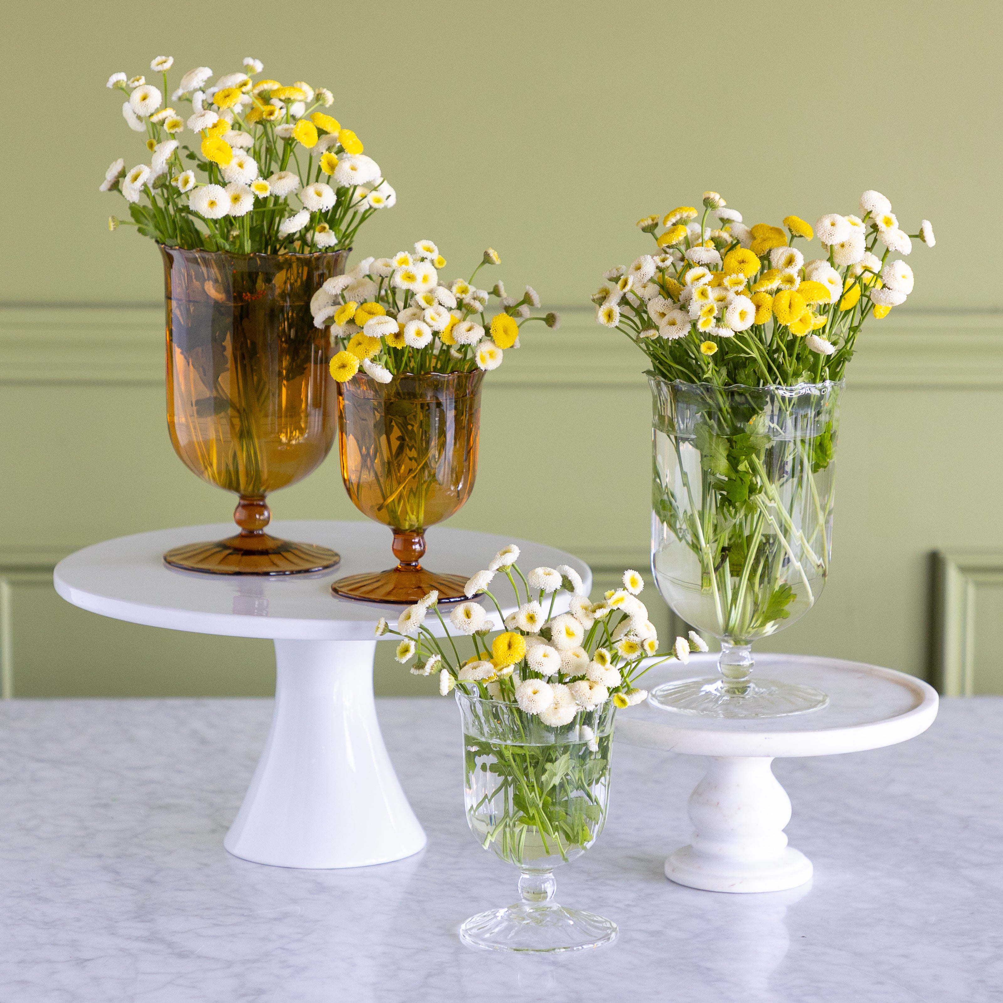 Two amber glass vase holders and two clear glass vase holders arranged on white pedestal stands with white and yellow daisy flower arrangements, set against a sage green paneled wall and light marble tabletop.