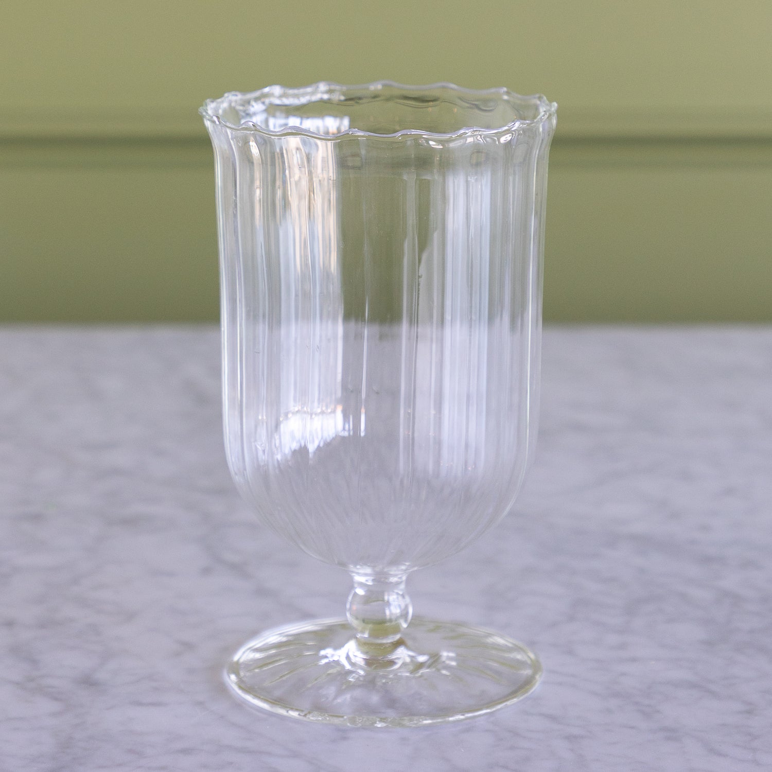 A clear ribbed glass goblet or candle holder with a scalloped rim and pedestal base, photographed on a light marble surface against a muted green wall.