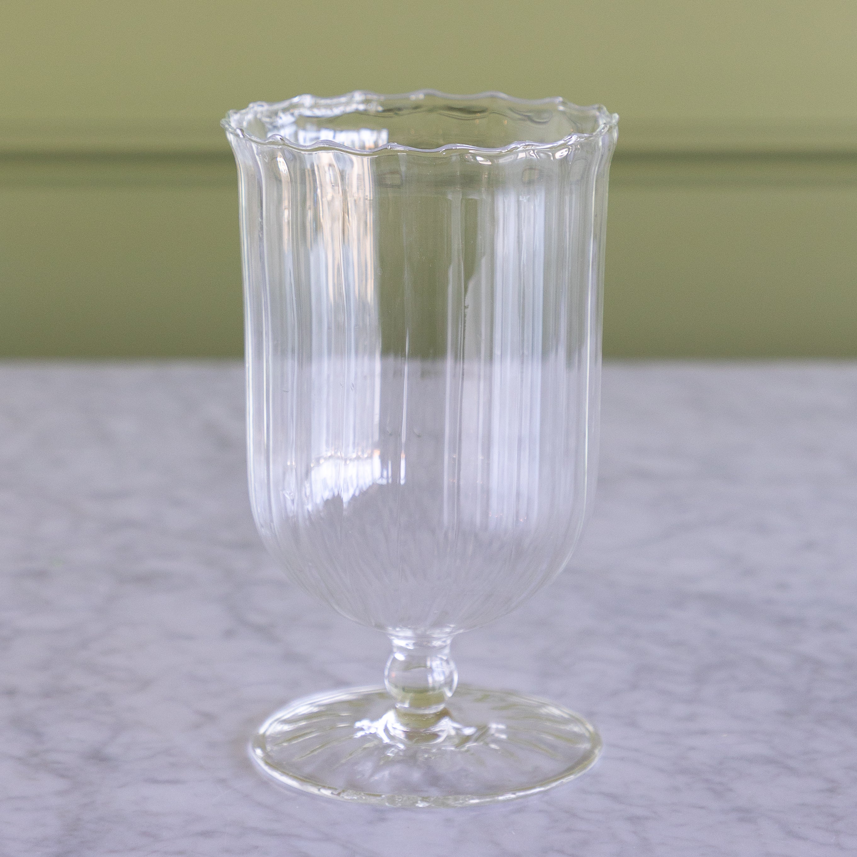 A clear ribbed glass goblet or candle holder with a scalloped rim and pedestal base, photographed on a light marble surface against a muted green wall.