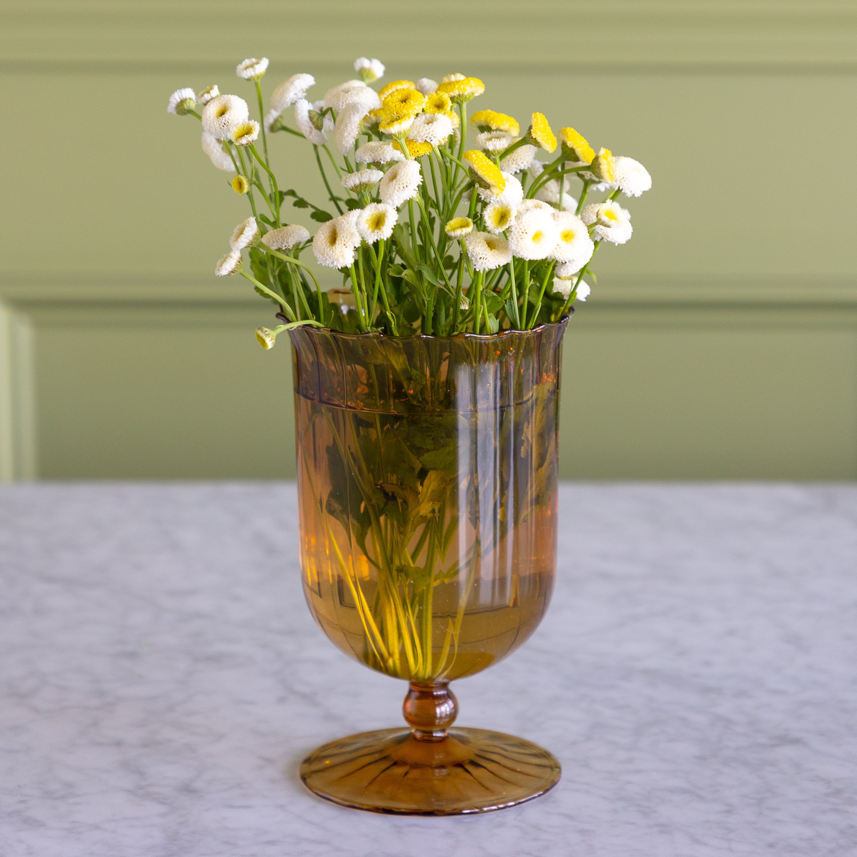 An amber glass goblet vase with a flared base, filled with white and yellow daisies, set on a light marble surface against a sage green paneled wall.