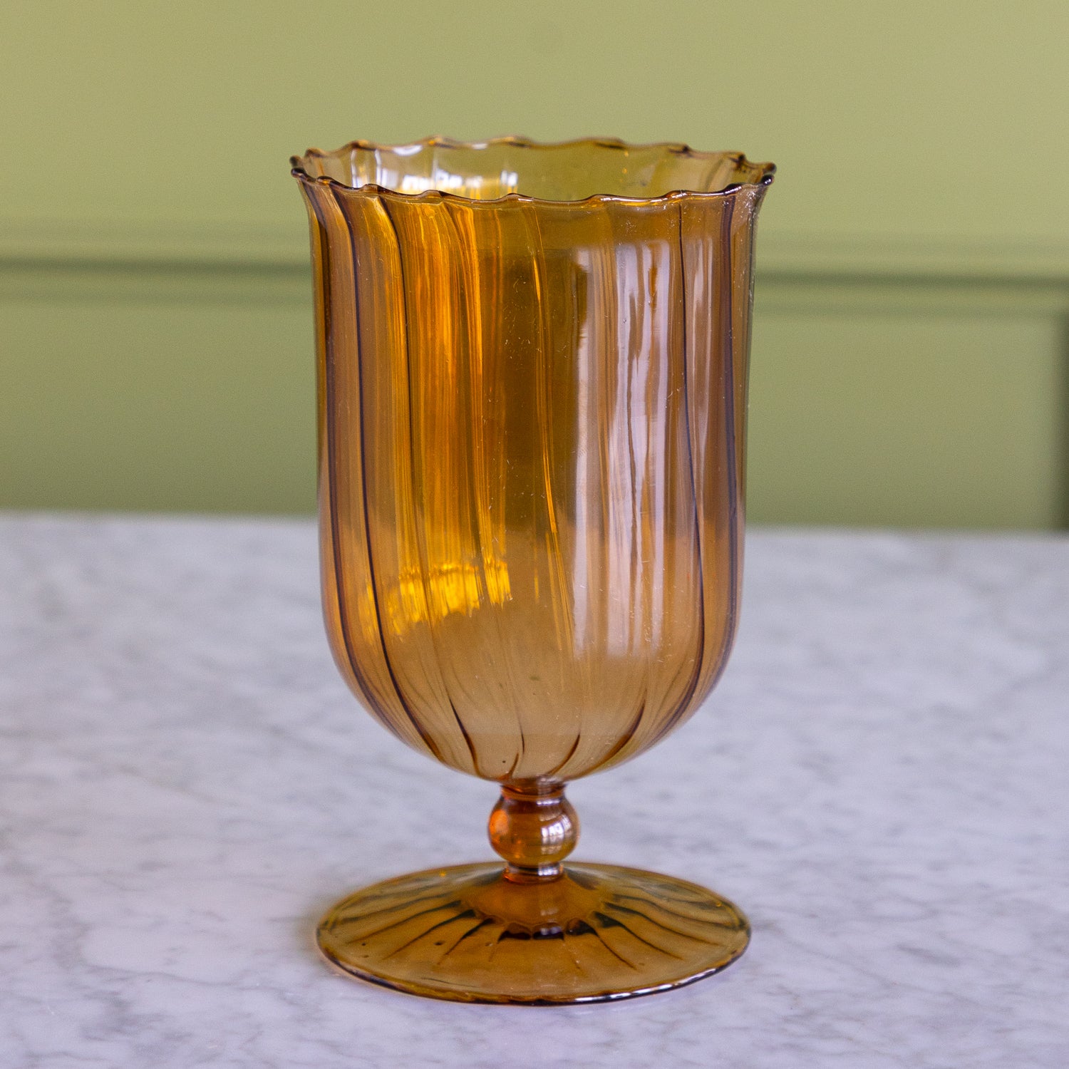 An amber-colored ribbed glass goblet with a scalloped rim and pedestal base, set on a white marble surface against a light green wall.