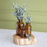 Three amber glass bottles of different sizes arranged on a round natural wood slice tray, with eucalyptus stems placed inside and around the bottles, set against a sage green paneled wall and white marble surface.