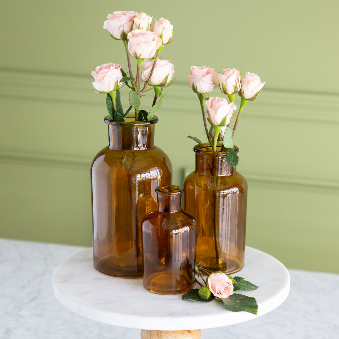 Three amber glass vases of different sizes arranged on a round white marble pedestal, each holding pale pink roses, against a light green paneled wall.