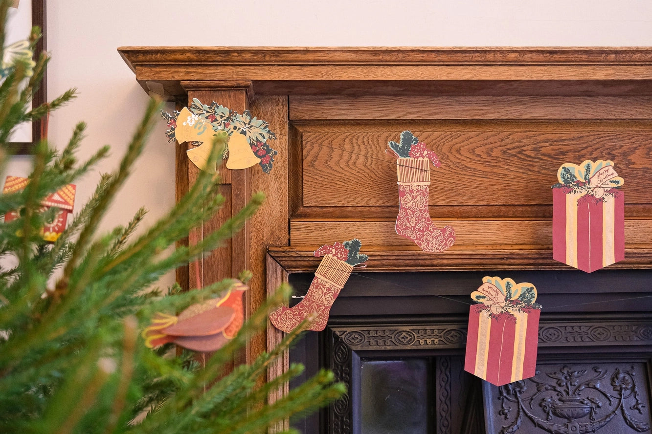 Paper Christmas mantel decorations including pink patterned stockings, gift boxes, a bird ornament, and a floral wreath, displayed on a dark fireplace with a blurred green pine branch in the foreground.