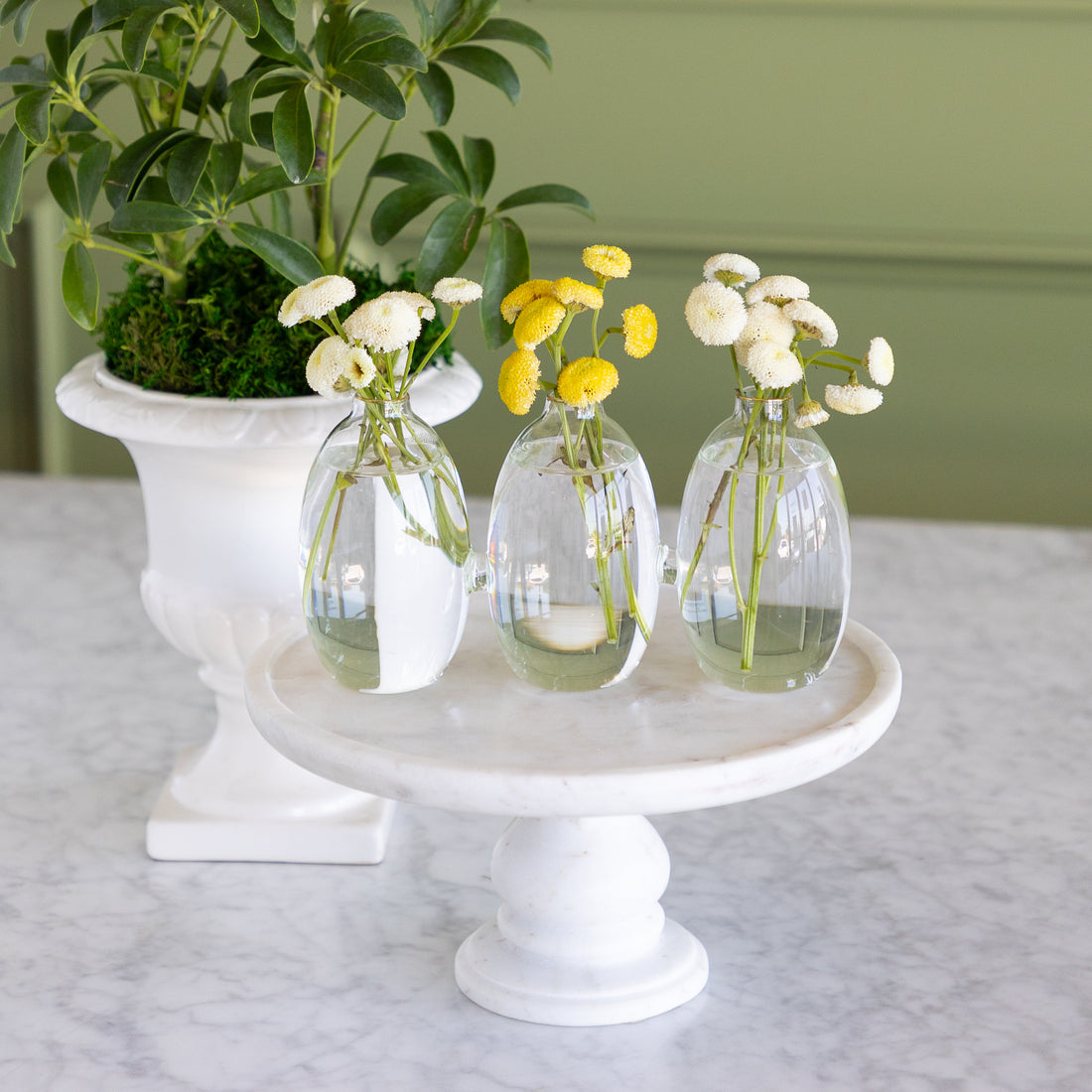 Vase with 3 cylindrical sections that are all attached together.  The vase has white and yellow small flowers in them on a white marble pedestal with a plant in the background. 