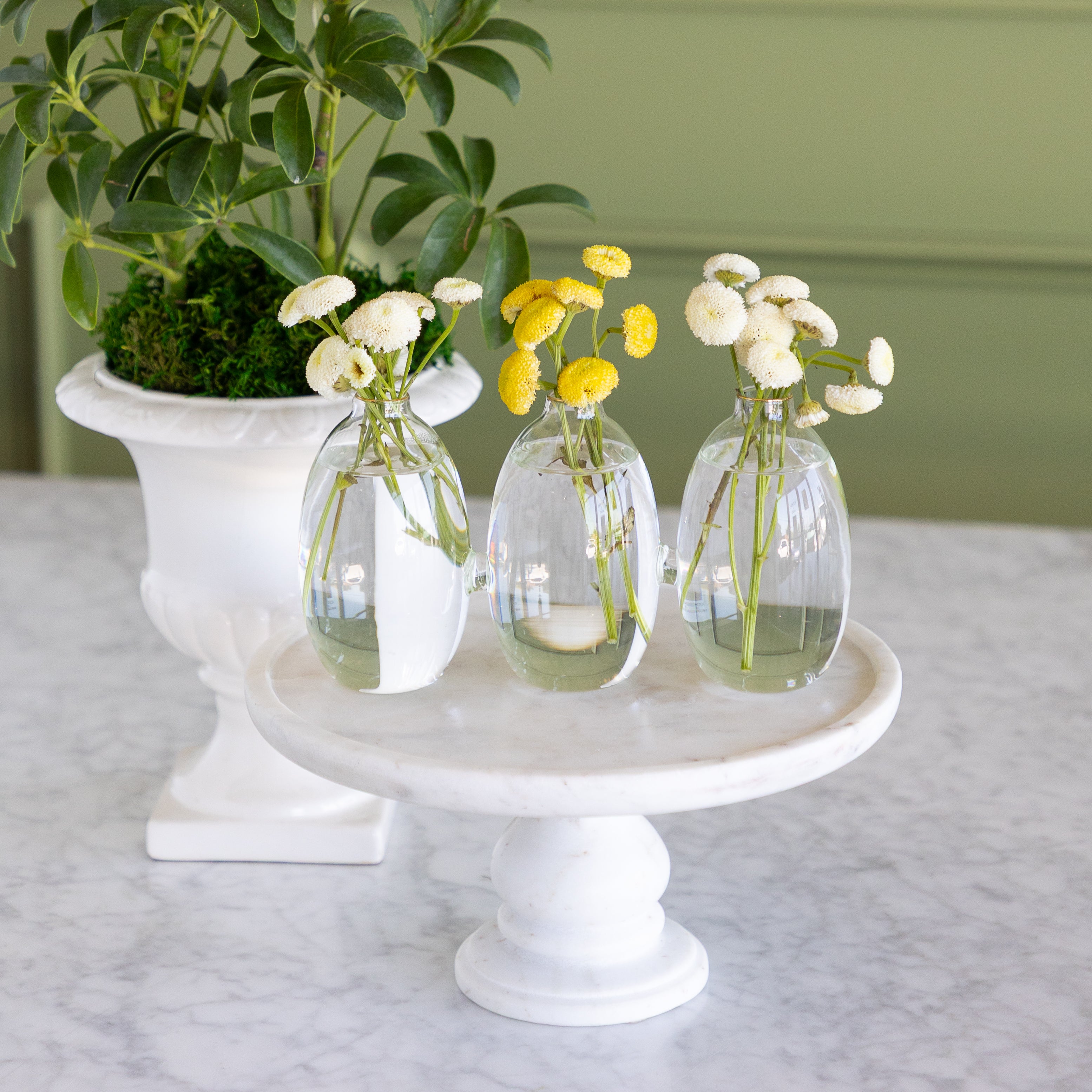 Vase with 3 cylindrical sections that are all attached together.  The vase has white and yellow small flowers in them on a white marble pedestal with a plant in the background. 