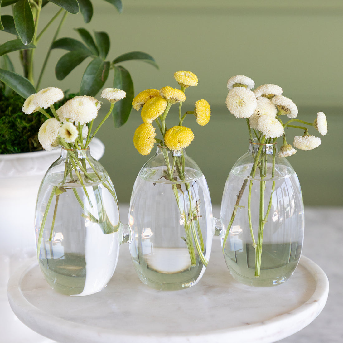 Vase with 3 cylindrical sections that are all attached together.  The vase has white and yellow small flowers in them on a white marble pedestal with a plant in the background. 