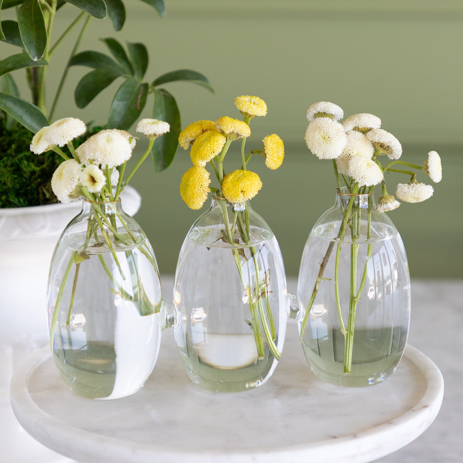 Vase with 3 cylindrical sections that are all attached together.  The vase has white and yellow small flowers in them on a white marble pedestal with a plant in the background. 