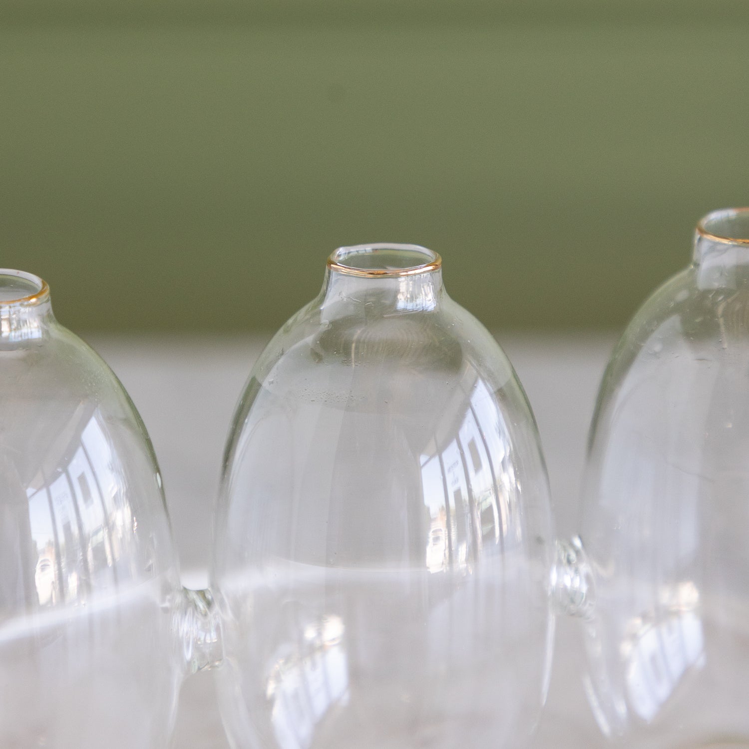 Three clear glass bud vases with small gold rims, arranged in a row against a soft green and white background.