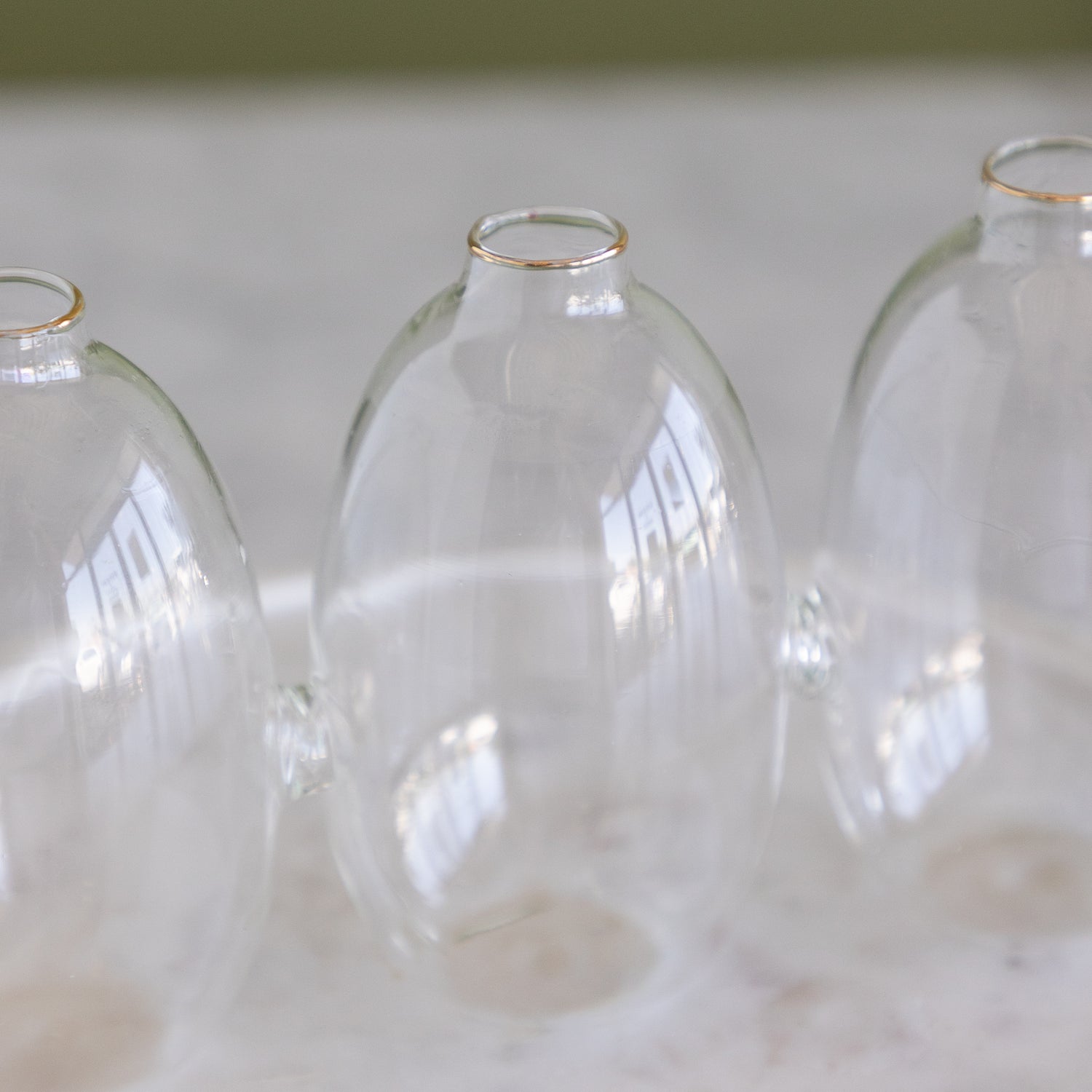 Three clear glass bud vases with small gold rims at the top, arranged on a light marble surface.