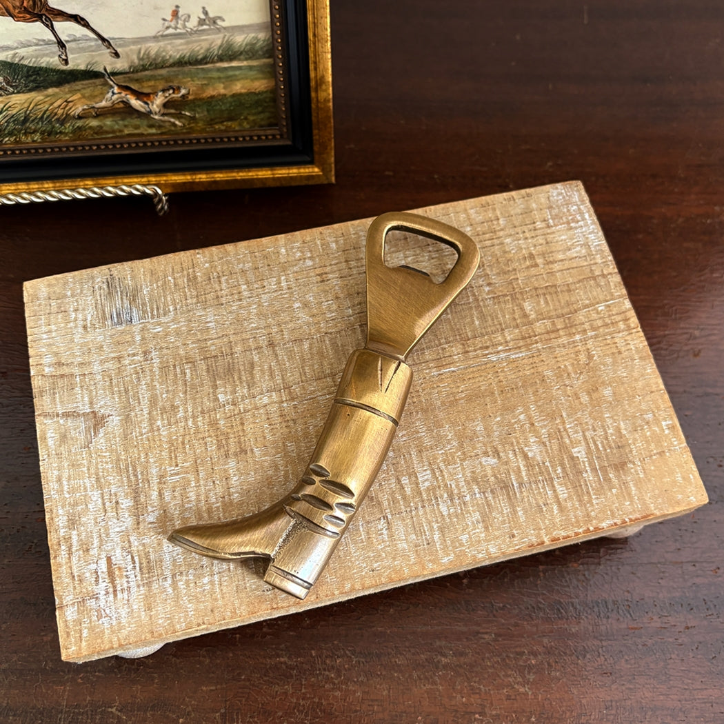 Brass boot-shaped bottle opener on a wooden surface with a painting in the background.