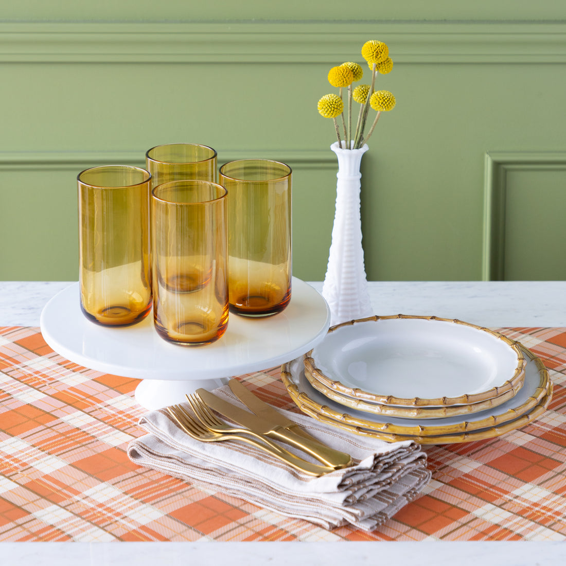 Table setting with amber glasses, gold cutlery, Juliska Bamboo Dinner Plates and a vase with yellow flowers on the Orange Plaid Runner.