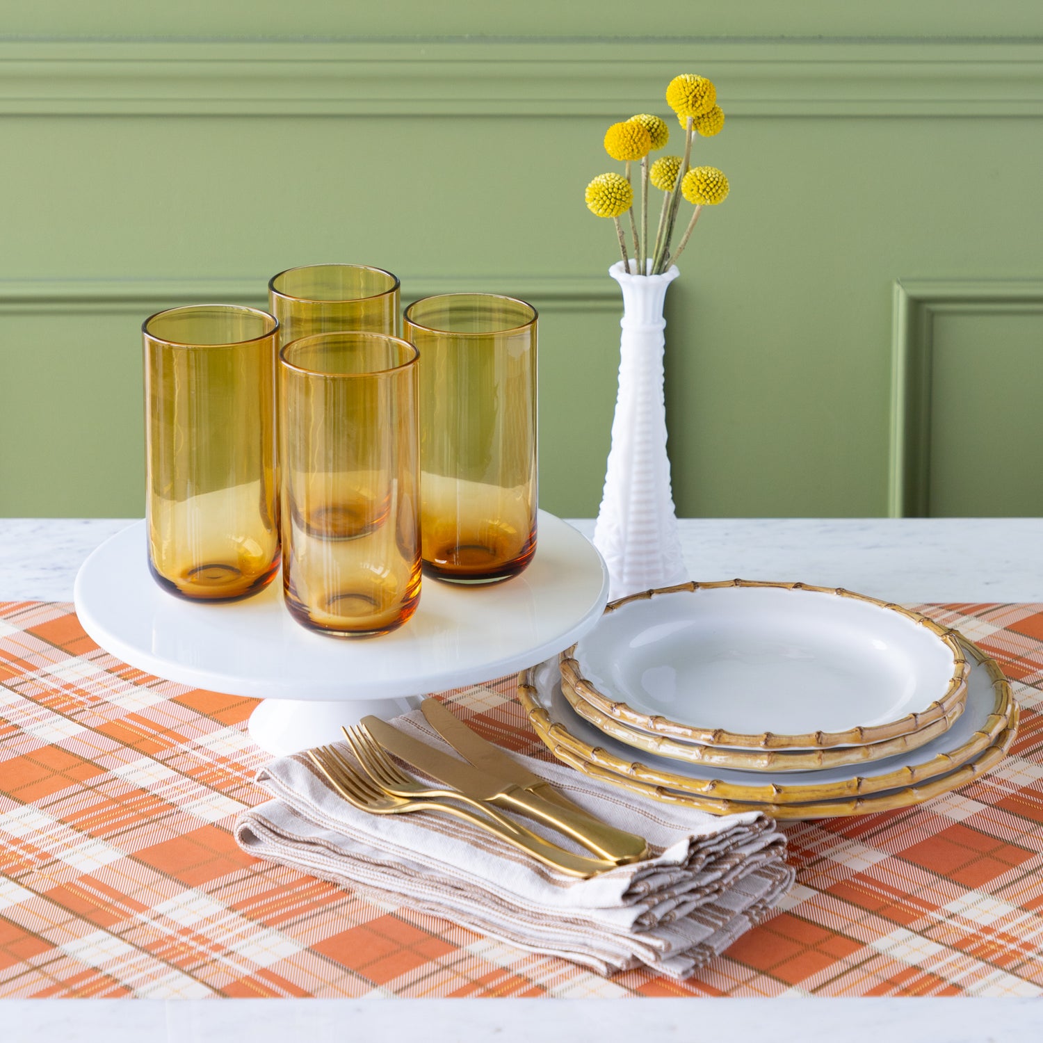 Table setting with amber glasses, gold cutlery, Juliska Bamboo Dinner Plates and a vase with yellow flowers on the Orange Plaid Runner.