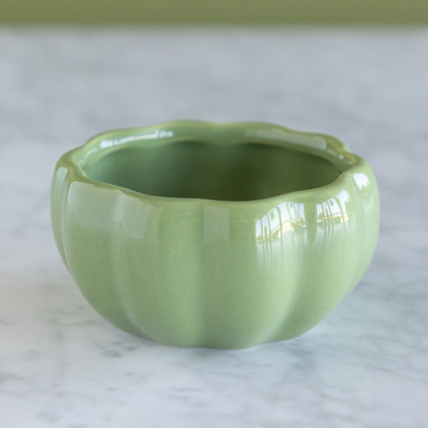 A small glossy ceramic bowl with a scalloped rim and ribbed body, shown on a light marble surface. The bowl is a soft light green color.