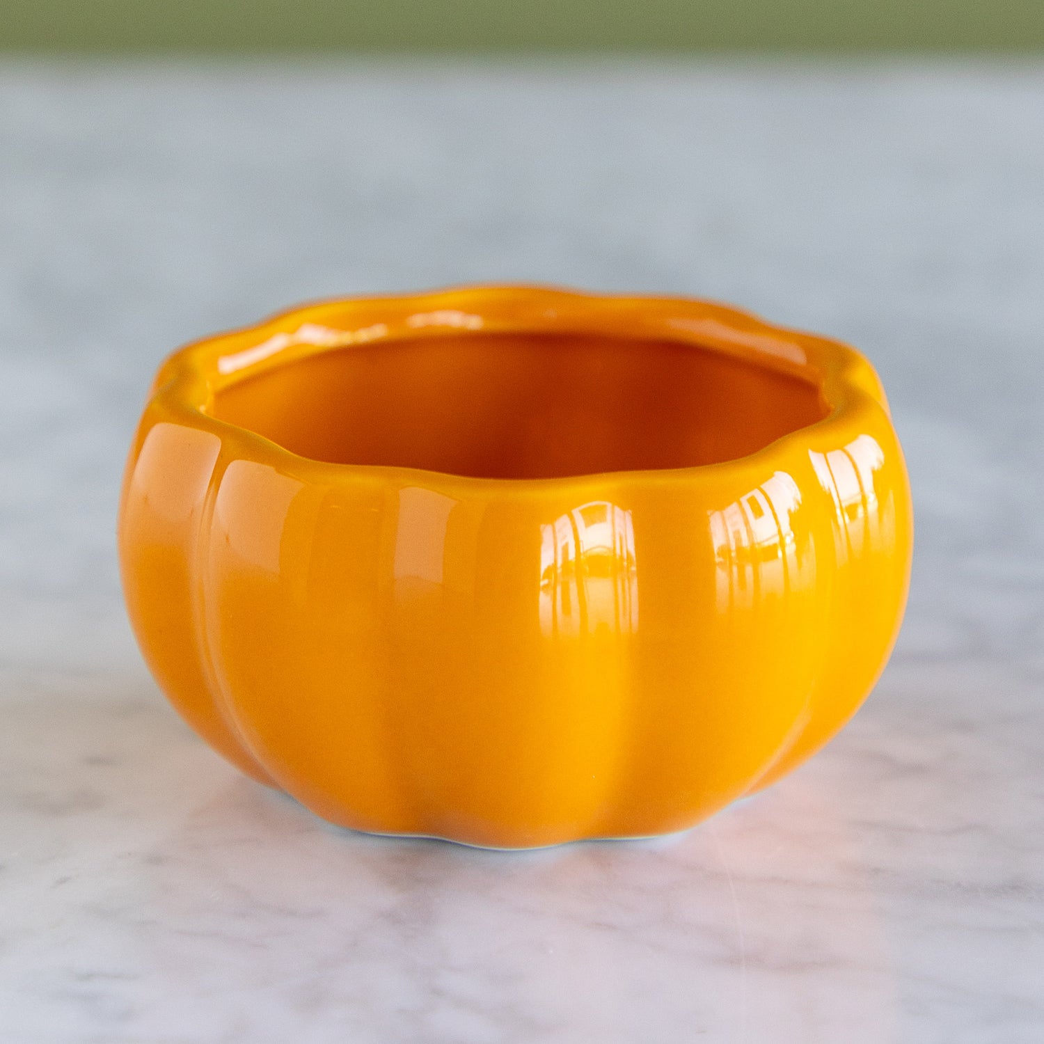 A glossy orange pumpkin-shaped ceramic planter or bowl with a ribbed design, sitting on a light marble surface.