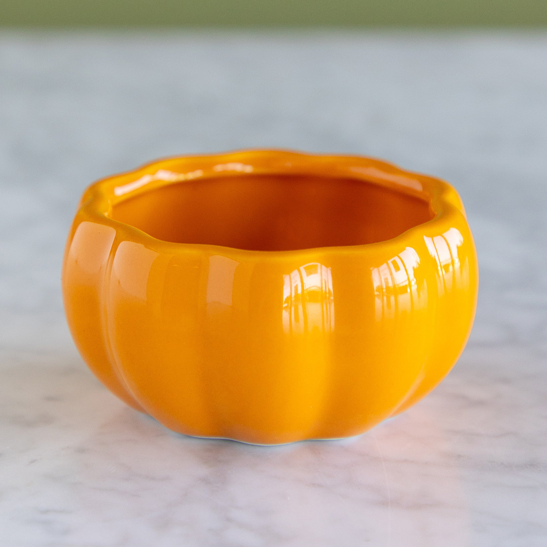 A glossy orange pumpkin-shaped ceramic planter or bowl with a ribbed design, sitting on a light marble surface.