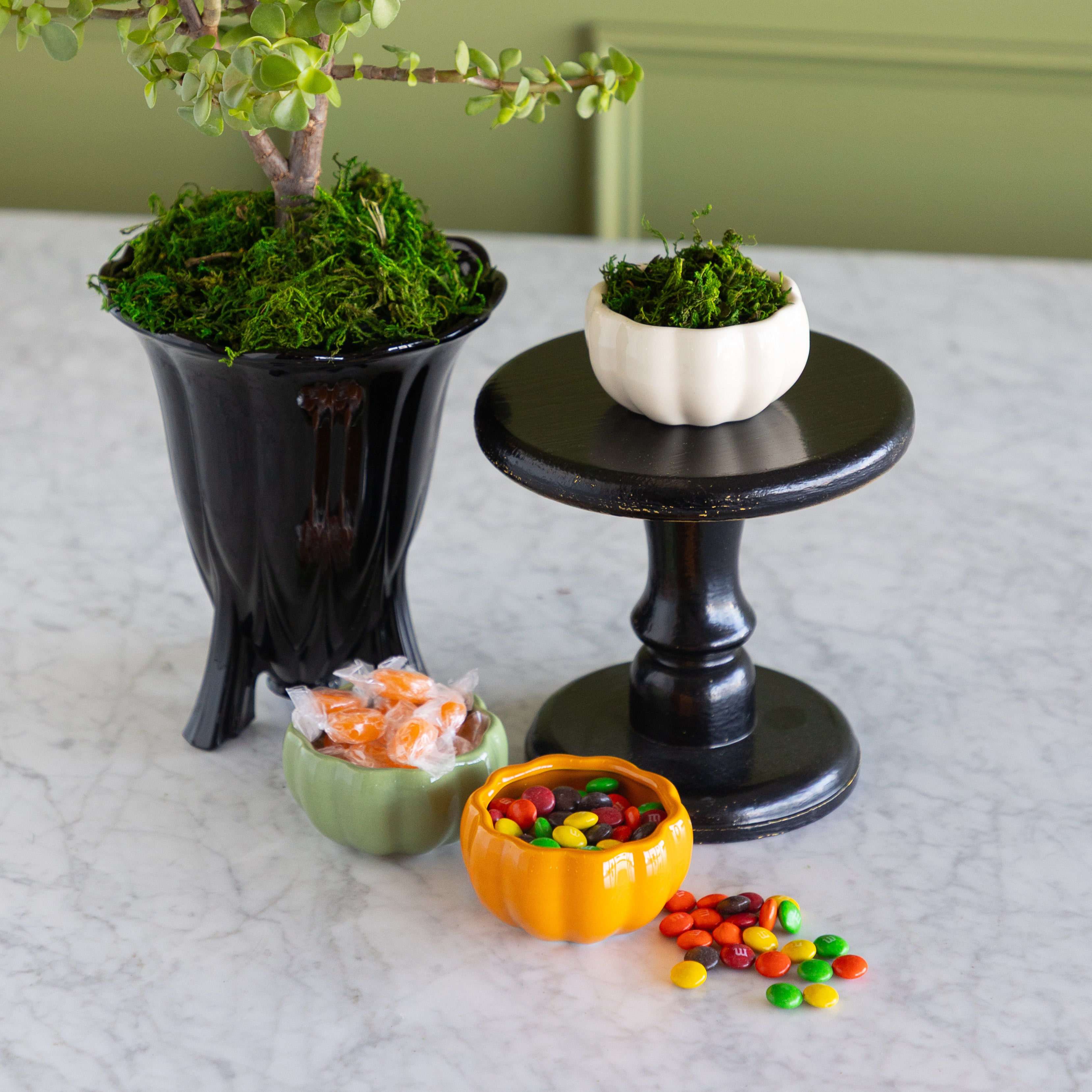A styled tabletop arrangement featuring a black pedestal stand holding a small white pumpkin bowl filled with green moss, alongside a large black planter with a tree and two small pumpkin-shaped candy bowls filled with colorful candies on a light marble surface.