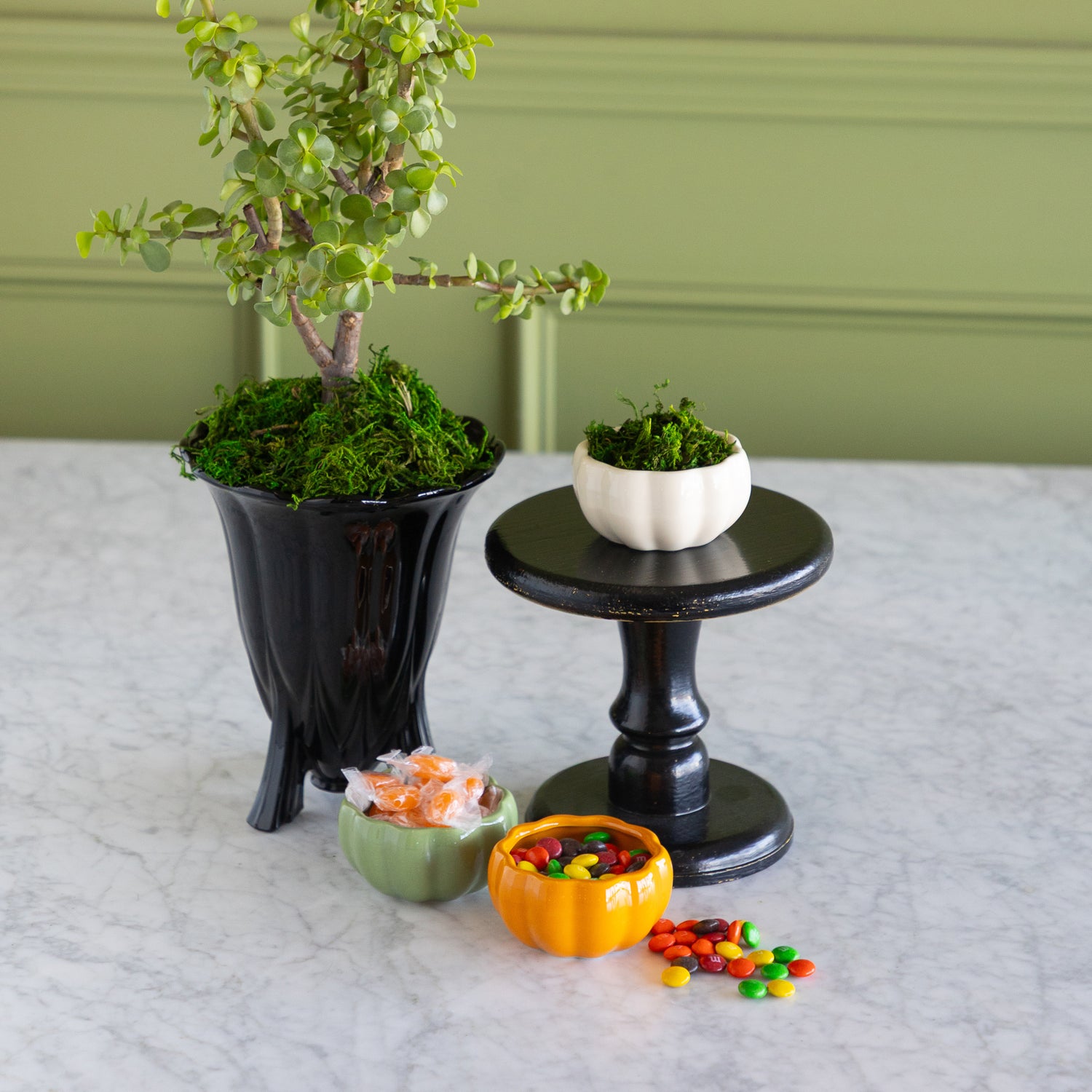 A styled tabletop arrangement featuring a potted green tree in a black planter, a small white pumpkin bowl of greenery on a black pedestal stand, and three pumpkin-shaped candy bowls containing candy on a light marble surface against a sage green paneled wall.