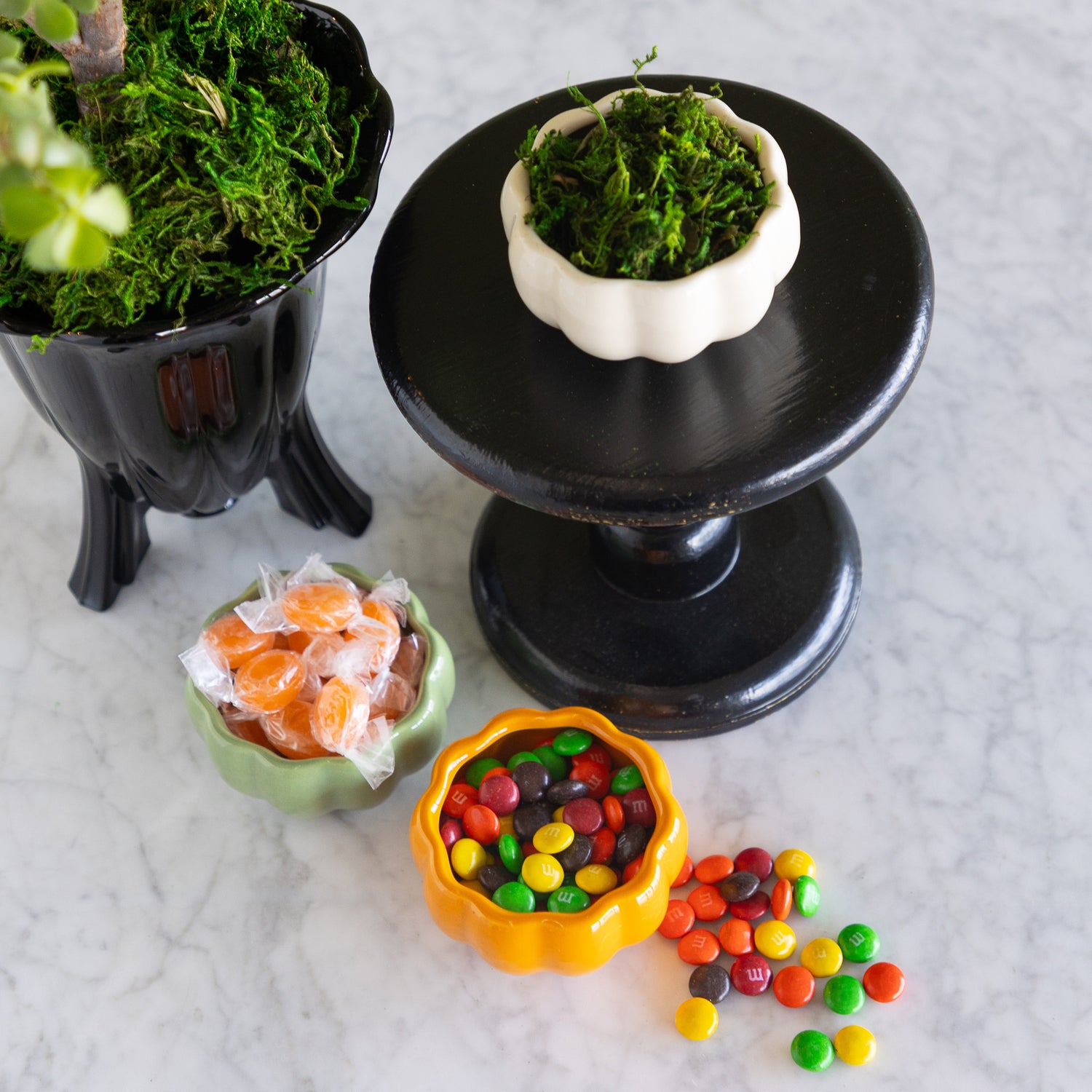 Three small decorative bowls filled with colorful candies and green moss, arranged on a light marble surface with a black pedestal stand behind them.