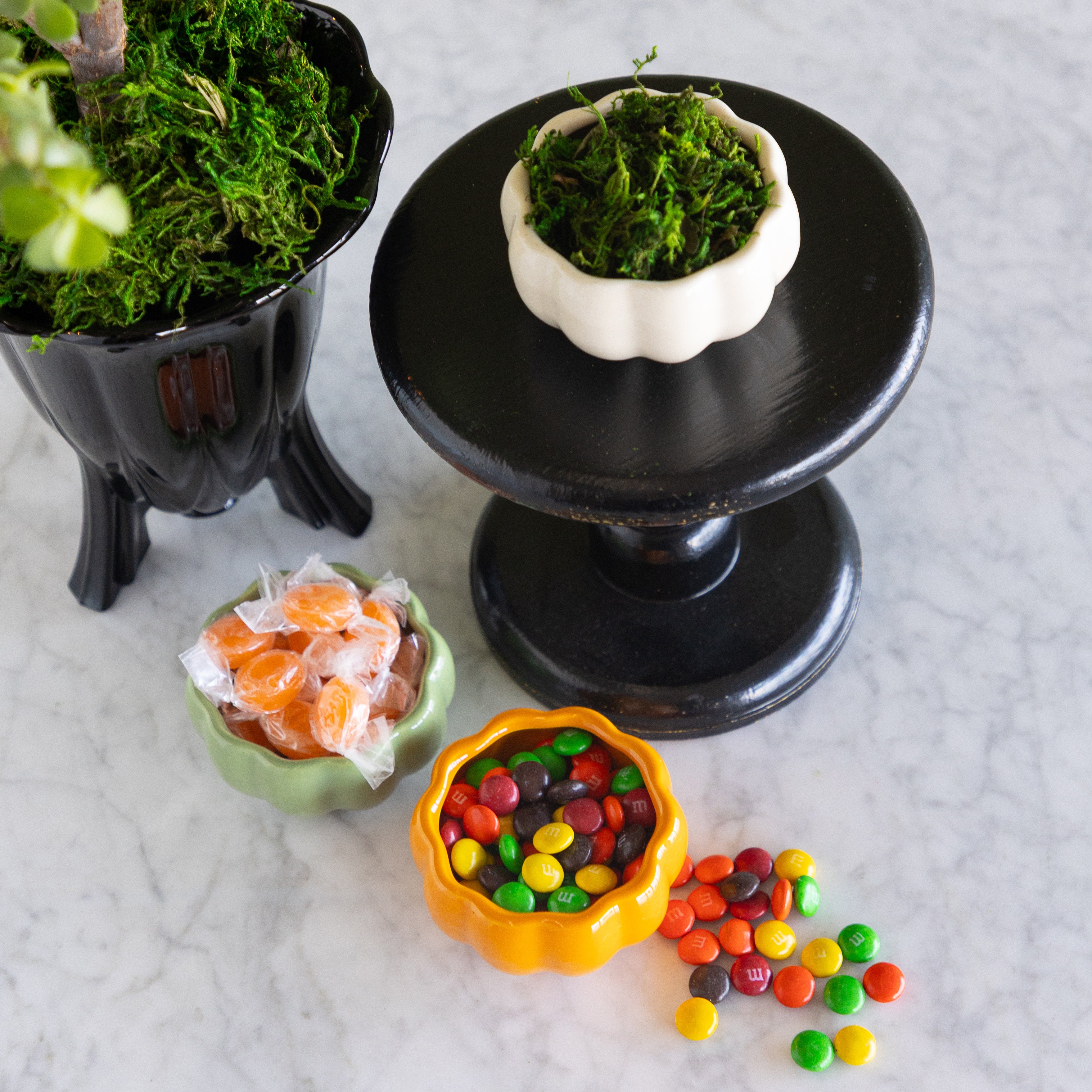 Three small decorative bowls filled with colorful candies and green moss, arranged on a light marble surface with a black pedestal stand behind them.