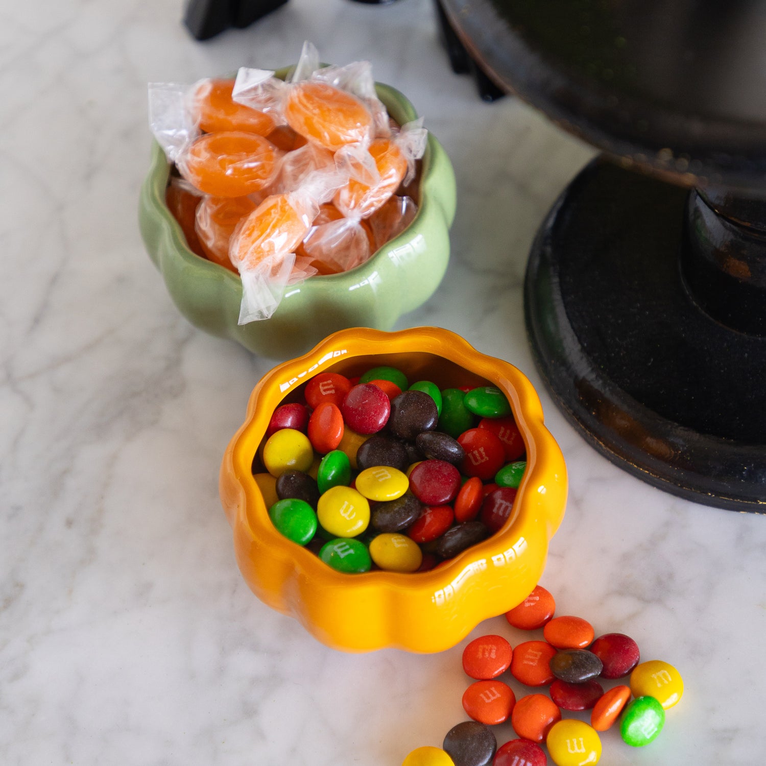 Two pumpkin-shaped bowls filled with candy on a white marble surface. The front bowl is orange and filled with multicolored M&amp;M&
