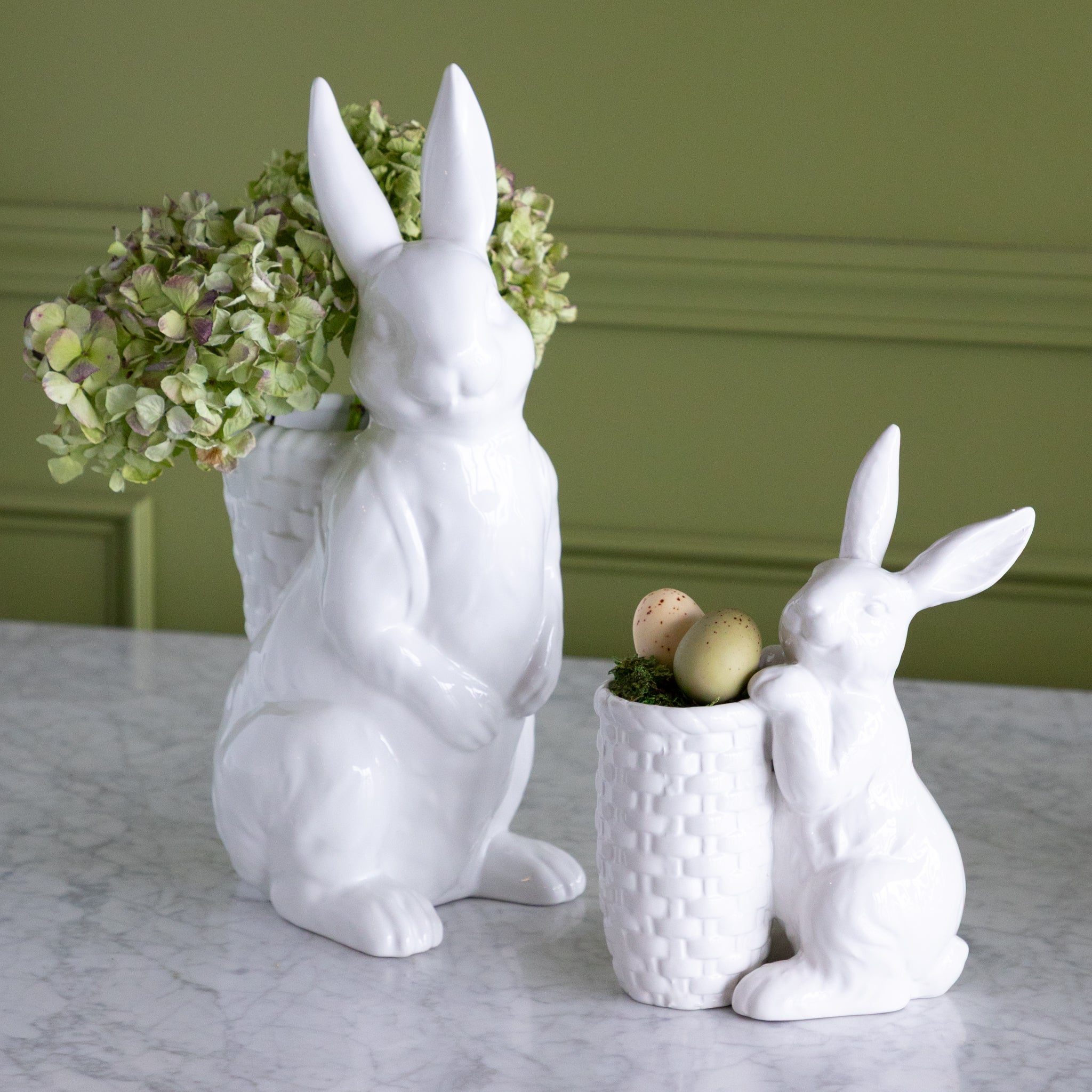 White rabbit-shaped planters with one holding green hydrangeas and the other with Easter eggs on a marble surface.