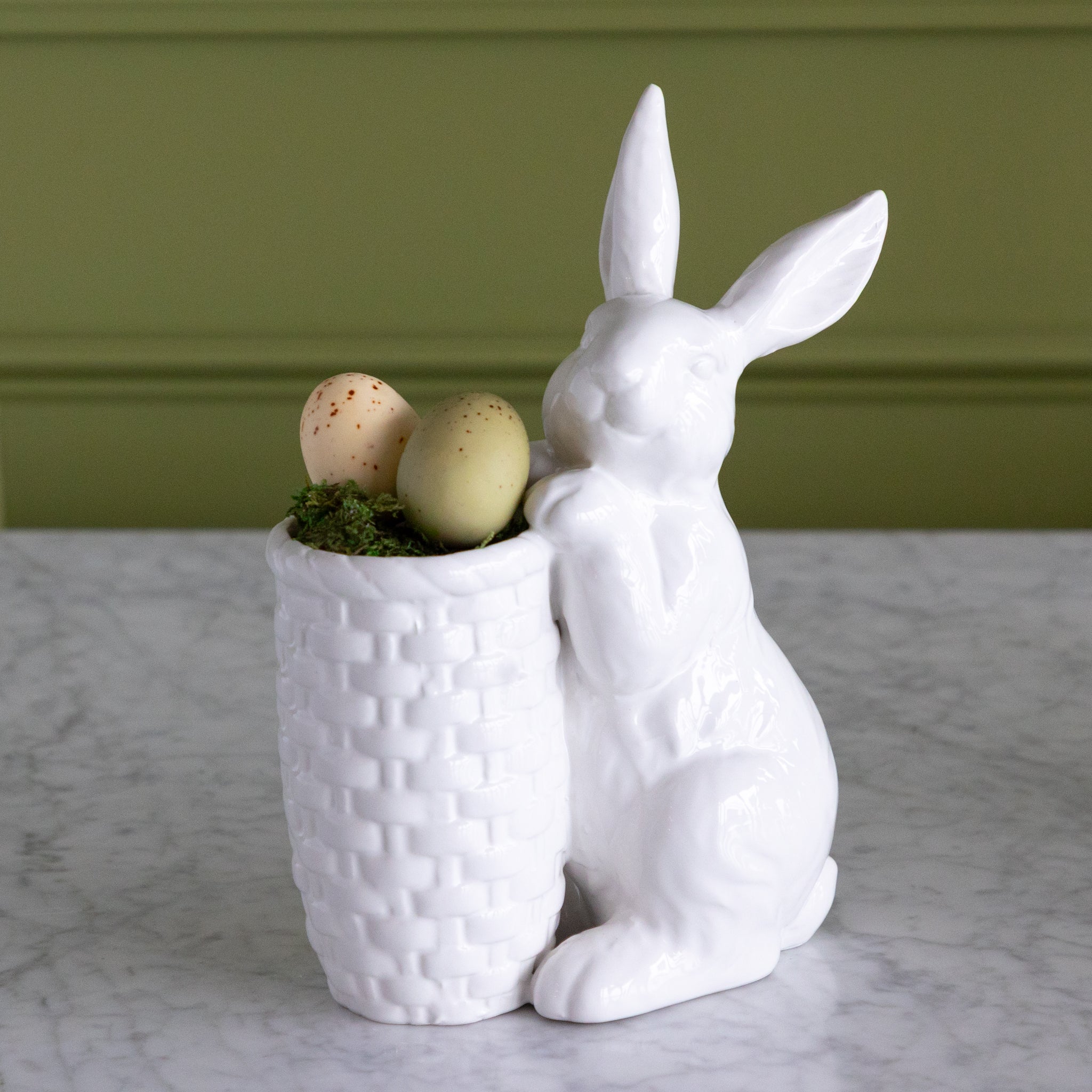 White ceramic rabbit holding a textured pot with Easter eggs against a green wall.