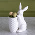 White ceramic rabbit holding a textured pot with Easter eggs against a green wall.