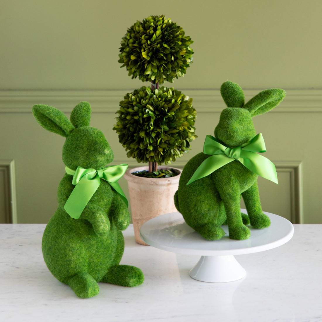 Two green moss-covered bunny figures with green ribbons and a topiary plant on a white surface.