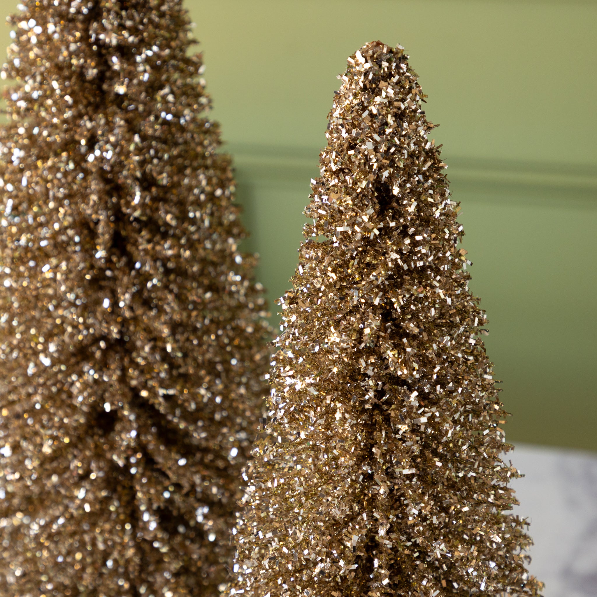 Two gold glittery bottlebrush trees on a marble surface with a green wall background.