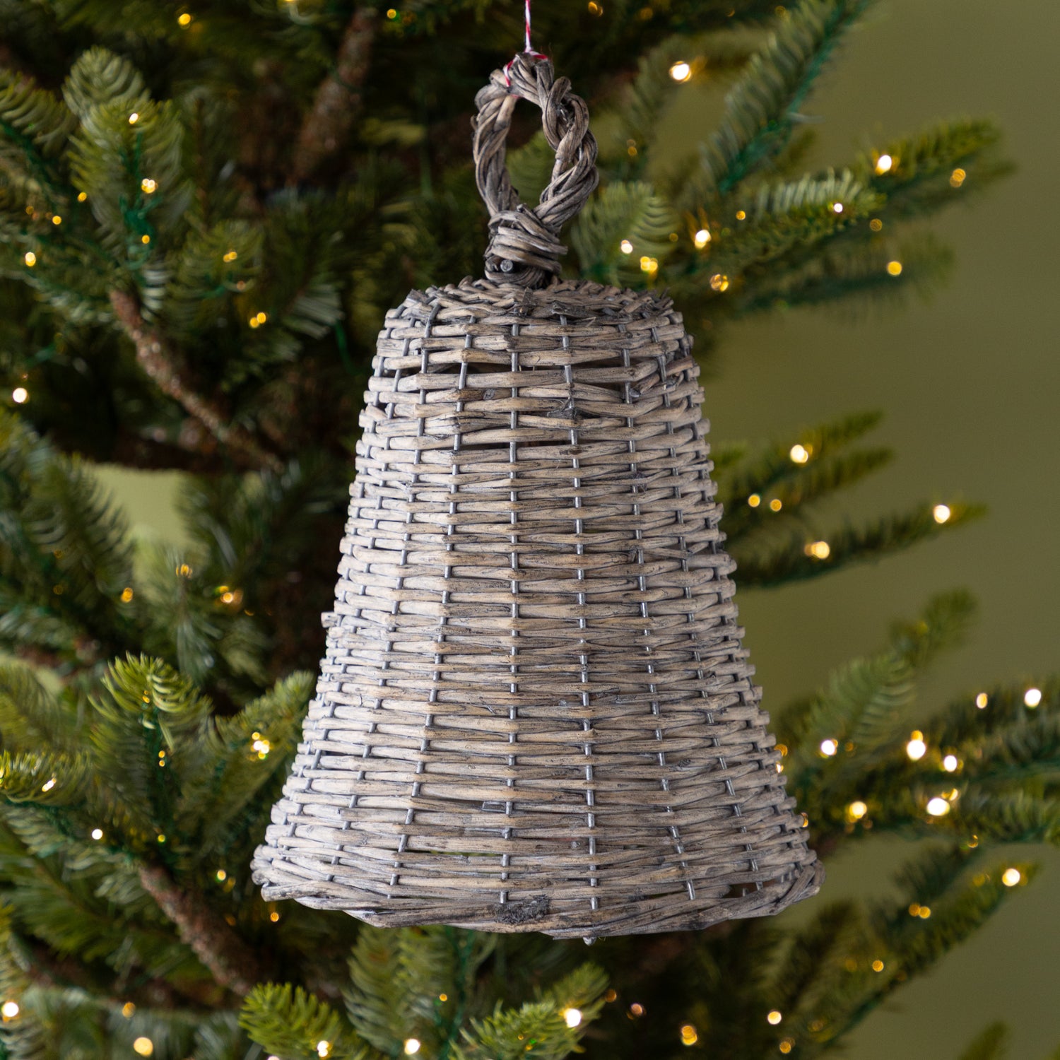 Wicker bell ornament hanging on a decorated Christmas tree with lights.