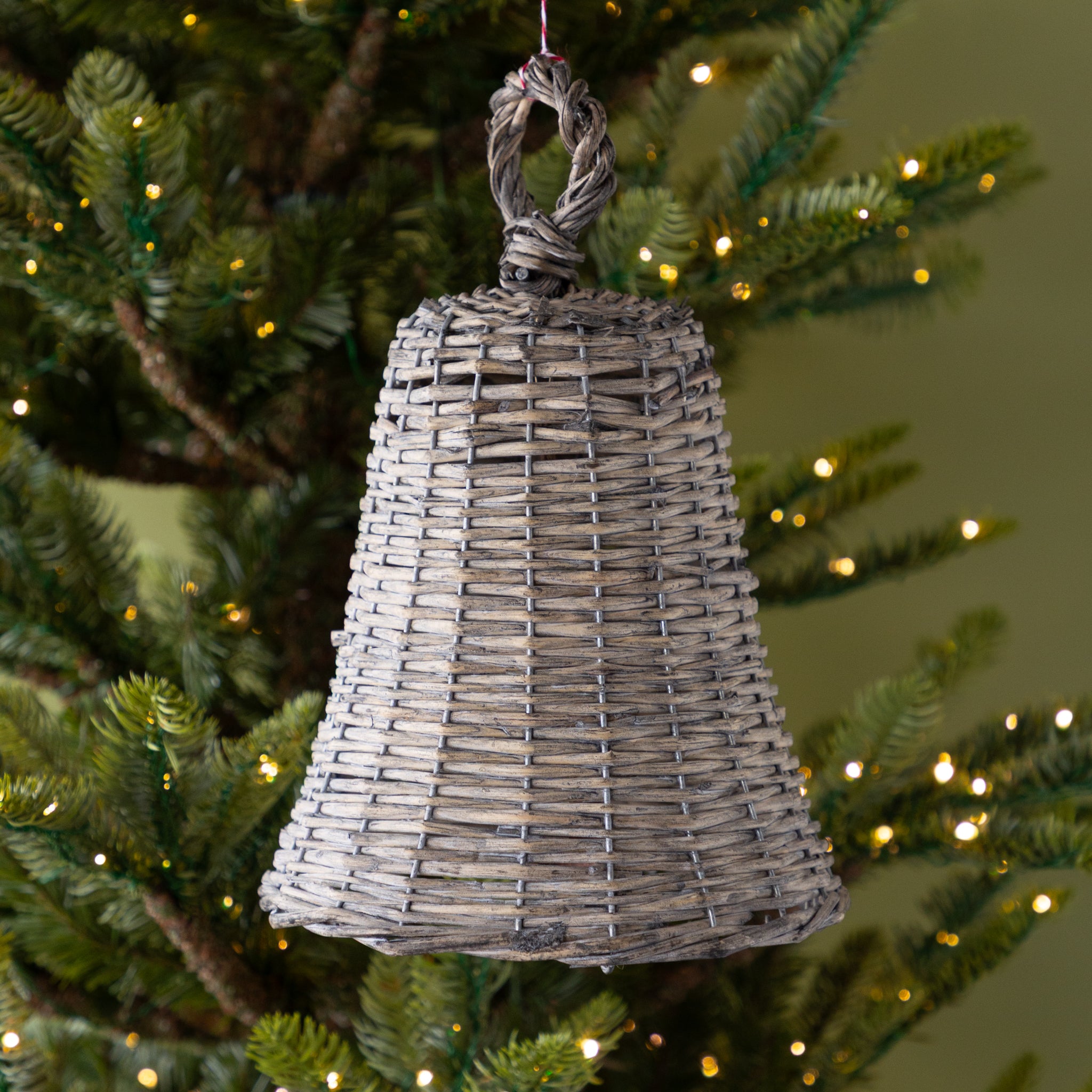 Wicker bell ornament hanging on a decorated Christmas tree with lights.