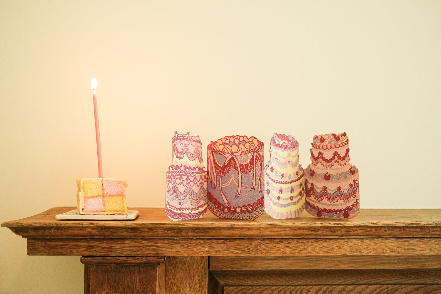 Concertina cake shaped greeting card on a mantle next to a birthday cake with a lit candle in it. 