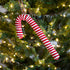 A red and white striped candy cane Christmas ornament hanging from a green Christmas tree with warm white lights.