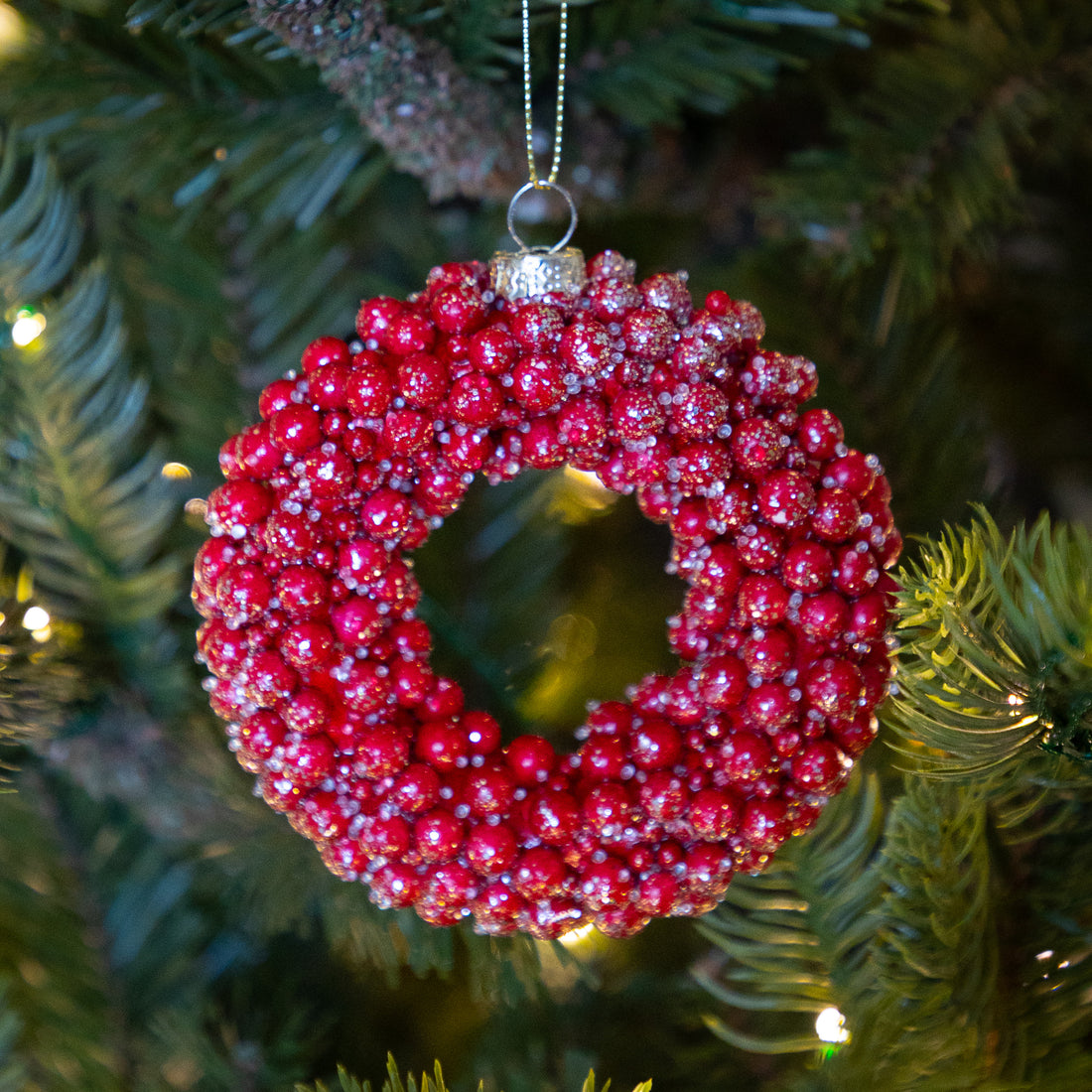 A red berry wreath Christmas ornament with silver glitter accents hanging from a silver chain on a green Christmas tree branch with warm string lights.