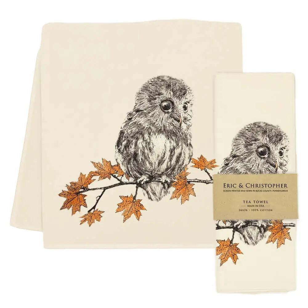 A cream-colored cotton tea towel with a detailed black-and-white owl perched on a branch with orange autumn leaves, shown alongside its paper packaging label.